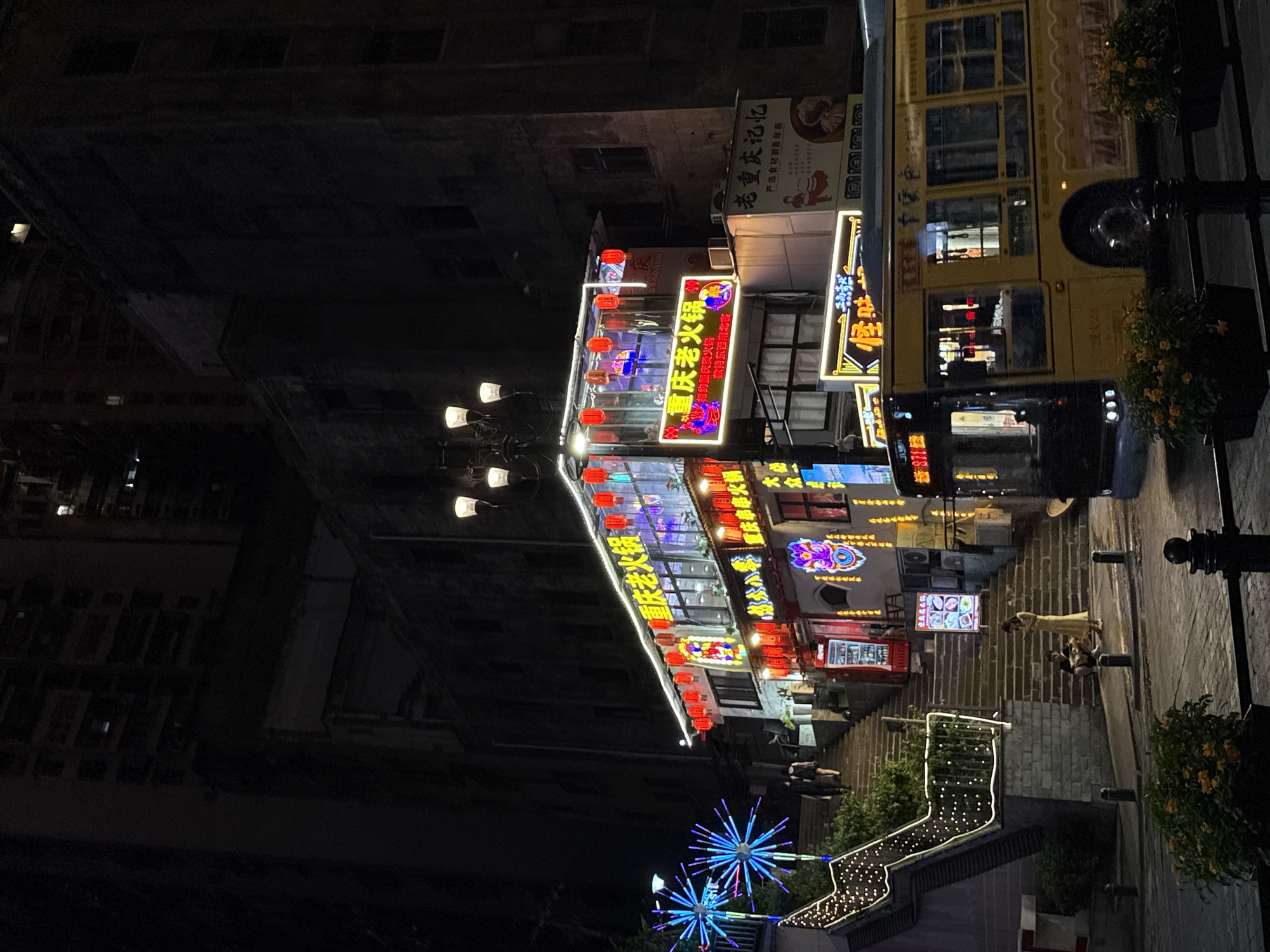 The image depicts a nighttime scene in the Yuzhong District of China, likely a commercial or entertainment area. A yellow bus is moving on the right side of the street, partially obscuring a building adorned with numerous red lanterns and neon signs. The signs are in Chinese characters, with one prominently displaying "重庆老火锅" (Chongqing Old Hot Pot). A set of illuminated stairs, decorated with string lights, ascends on the left side of the frame. Two individuals are walking up the stairs. Further down the stairs, a woman in a light-colored dress is standing, and a man is seated. The background consists of tall, dark buildings, typical of an urban setting at night.