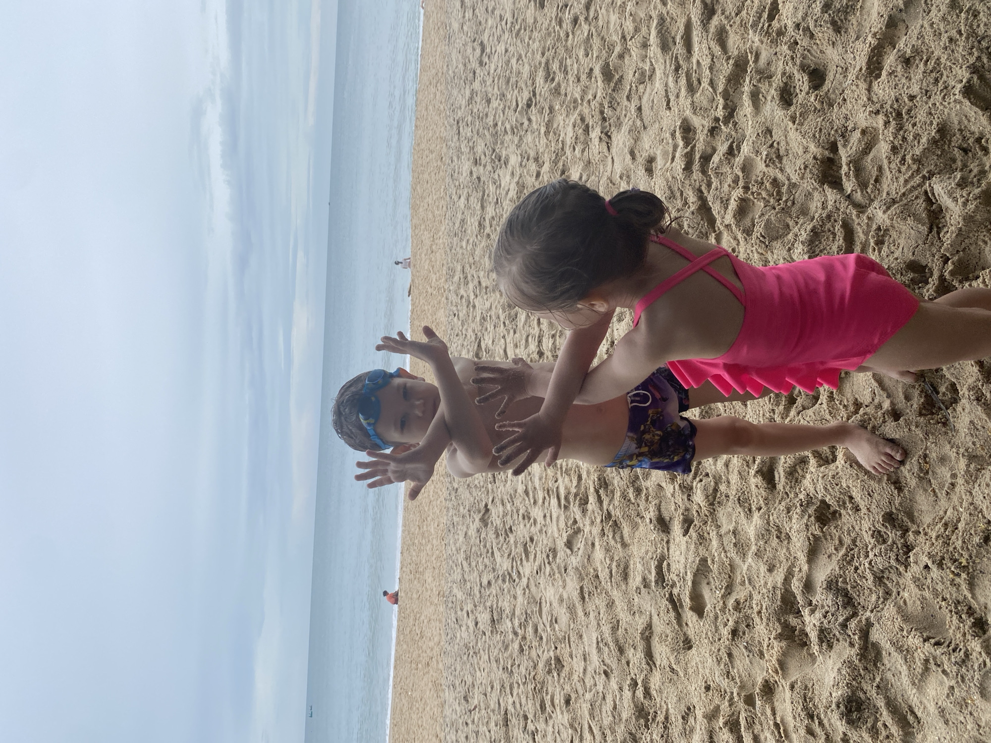 Two children are playing on a sandy beach in Phu Thuy Ward, Vietnam. The boy, wearing swimming goggles and patterned swimming trunks, is lying on his back with his arms raised. The girl, dressed in a pink swimsuit, is kneeling next to him, seemingly interacting with him. The background features the ocean and a cloudy sky. Other people are visible in the distance along the shoreline. The beach surface shows footprints in the sand.