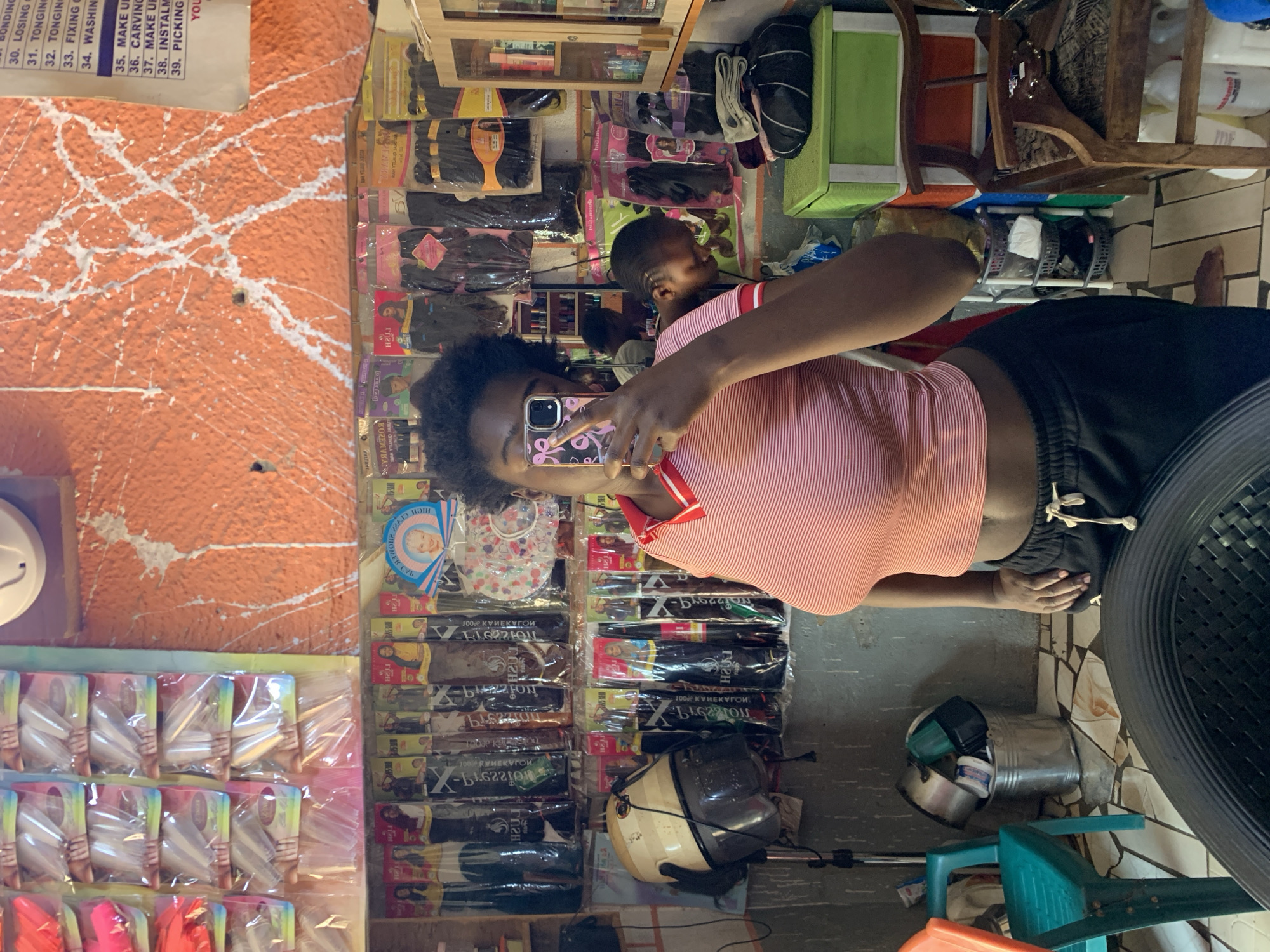 This is a selfie taken in a hair salon in Rafin Par, Nigeria. The image shows a woman taking a picture of herself in a mirror. She is wearing a pink and white striped crop top and black sweatpants. In the background, there are shelves stocked with hair products, including braids, weaves, and hair extensions. There is also a salon dryer visible. The walls are painted orange with white paint splatters, and there is a sign with a list of hair services. A young boy can be seen in the background. The floor is tiled.