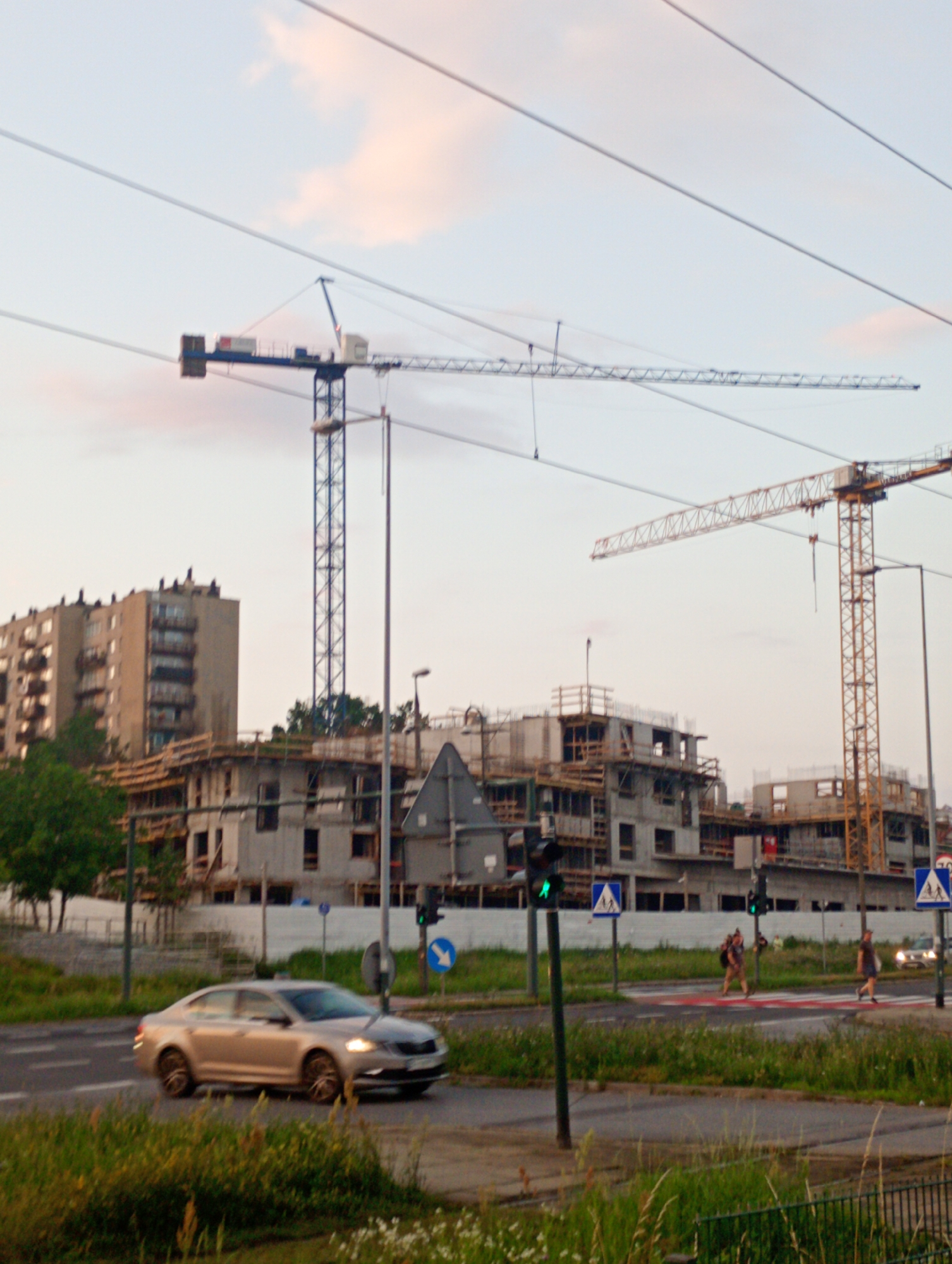 The image depicts a construction site in Krakow, Poland, featuring two prominent tower cranes against a pale sky with wispy clouds. In the foreground, a silver sedan is captured in motion on a paved road. The construction site itself shows the early stages of building, with visible concrete structures and scaffolding. Two individuals are seen walking across a pedestrian crossing, marked with red and white stripes. Traffic lights are present, one of which is illuminated green, indicating a favorable signal. The surrounding environment includes grassy areas and trees, with a multi-story apartment building visible in the background to the left. The overall scene suggests an active urban development area.