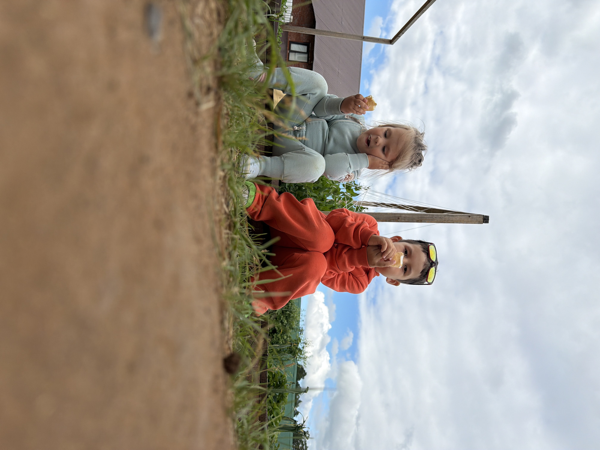 Two children, a girl and a boy, sit outdoors on a wooden surface. The girl, on the left, wears a light blue tracksuit and has her hand on her cheek as she eats. The boy, on the right, wears an orange tracksuit and sunglasses pushed up on his head, and he is also eating. They are surrounded by grass and dirt, with a building and greenery in the background under a cloudy sky.