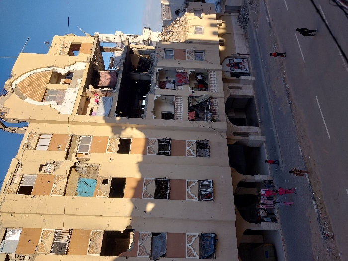 This image depicts a multi-story residential building in Khan Yunis, Palestinian Territory, presenting extensive structural damage. The upper right portion of the building, specifically the top two floors, exhibits severe destruction, including a large arched section that appears to have been blown out. Numerous windows throughout the building are shattered, missing, or obscured; a blue tarpaulin covers an opening on the third floor from the bottom on the left side. The facade shows widespread pockmarks, cracks, and discolored sections consistent with battle damage or shelling. Despite the damage, laundry is visible hanging on several balconies, indicating continued occupancy in parts of the building.
At the ground level, a paved street with white lane markings spans the foreground. A sidewalk, constructed from lighter grey paving blocks, runs parallel to the building. Under the building's arched ground-floor overhang, a group of approximately ten individuals, mostly children, are gathered on the left side of the sidewalk. A light-colored car is partially visible parked beneath these arches on the far left.
Several other individuals are present on the street and sidewalk: a child in a red shirt is on the sidewalk to the right of the main group; another child in a patterned shirt walks on the street in the left foreground; a child in a red shirt walks on the street in the middle-right; and a person in dark clothing walks on the street in the right foreground. A pile of rubble is visible on the ground to the far right, adjacent to the damaged building. A partially obscured mural, featuring a red character, is visible on a wall under the building's arches on the right. The sky is clear blue, and long shadows suggest either early morning or late afternoon lighting.
