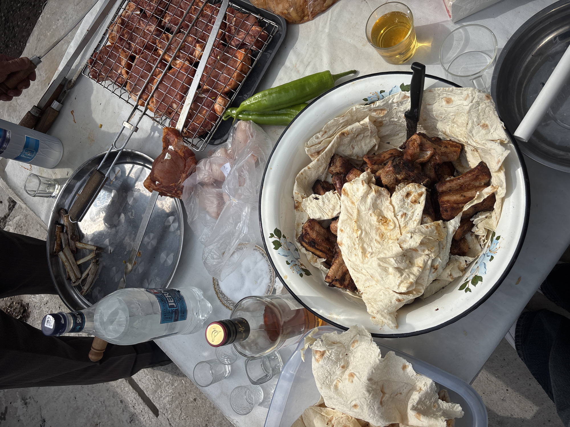 The image depicts an outdoor dining setup in Abovyan, Armenia. A low white table is laden with food and drinks. On the left, meat is grilling in a metal cage, next to metal skewers holding cooked meat. A metal tray contains remnants of grilled meat. A large white bowl, decorated with blue flowers, is filled with grilled meat and lavash bread. Nearby are two green peppers, a salt dish, and a clear plastic bag containing unknown contents. Drinks include a glass of amber liquid, likely alcohol, and two bottles of clear liquid, one labeled "JERMUK" in Armenian. Several small glasses sit near the bottles. Lavash bread is also piled in a clear plastic container. Part of a person is visible to the left and right. The setting appears to be sunny and outdoors.