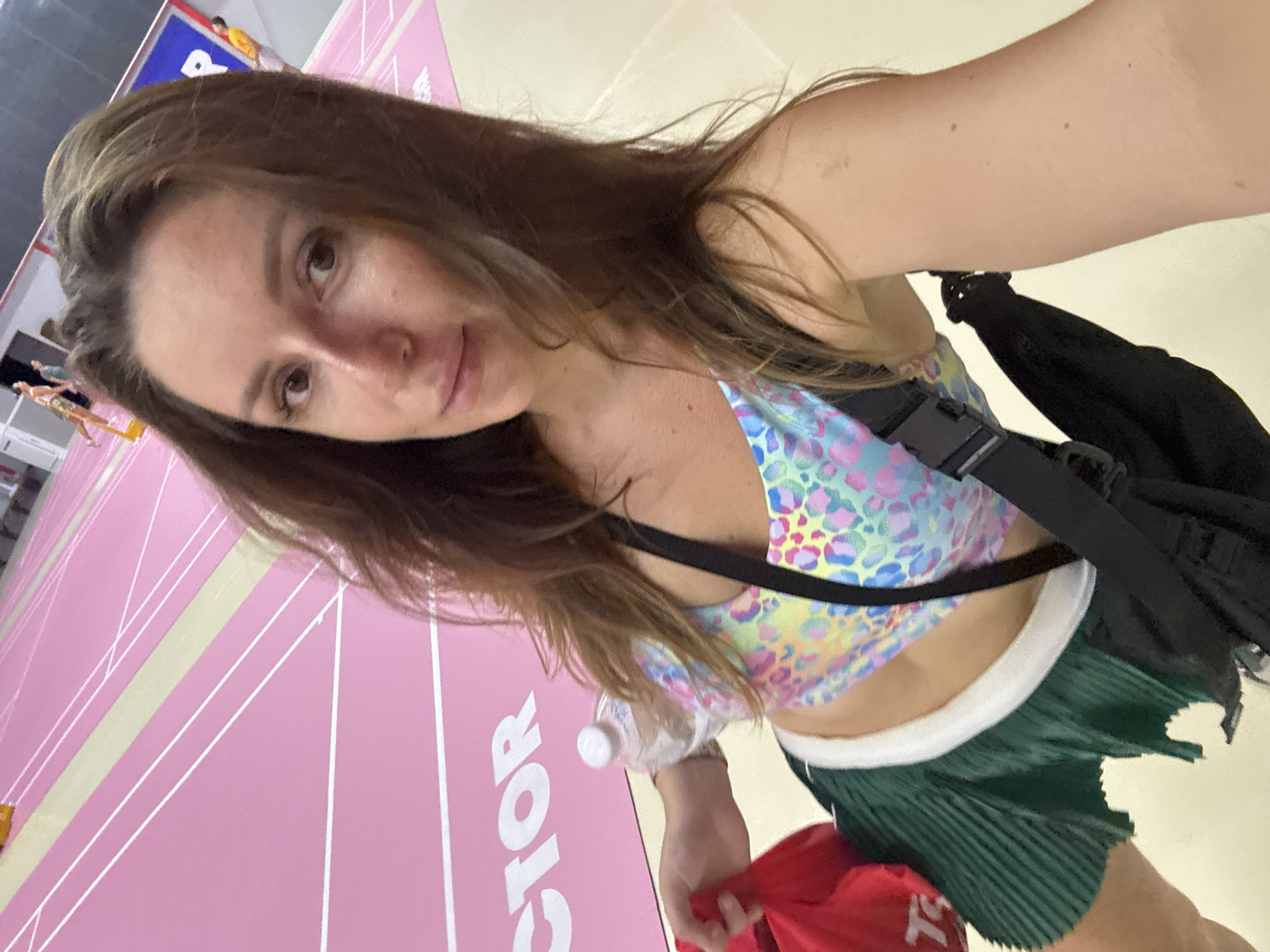 A woman is taking a selfie in an indoor sports facility with a pink track. She is wearing a colorful leopard print sports bra, a green pleated skirt, and has a black bag slung over her shoulder. In her left hand, she is holding a red bag and a water bottle is visible in the background. Other people are in the background on the track.