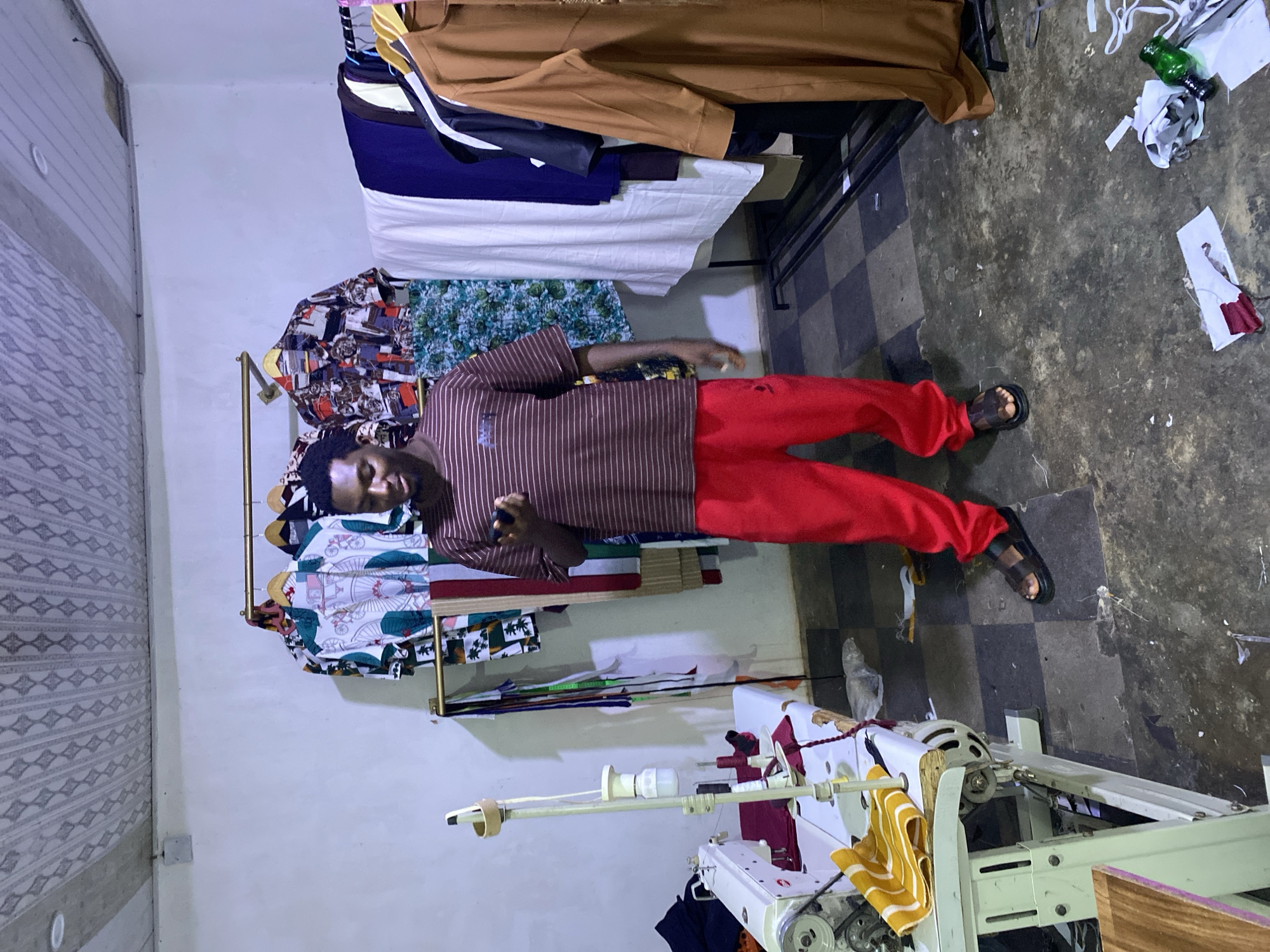 The image shows an indoor scene, likely a clothing workshop or small retail space. A man with dark skin and short dark hair stands centrally, looking directly at the camera with a neutral expression. He is wearing a brown short-sleeved shirt with thin vertical white stripes, bright red trousers, and dark sandals. He holds a small dark object in his right hand.
Behind the man, on his left, a clothing rack displays several patterned and colorful shirts. Further back, another display or table holds neatly folded fabrics and garments, including brown, dark blue, and white items. To the man's right, an industrial-style sewing machine is partially visible in the foreground, with a yellow and white striped cloth draped over part of it.
The walls are light-colored, with a patterned design on the wall to the left. The floor features a grey and black checkered pattern in some areas, transitioning to a darker, more textured concrete-like surface. Fabric scraps, paper, and a green bottle are scattered on the floor, suggesting an active workspace. The lighting appears to be artificial, typical of an indoor setting. There is no visible, decipherable text in the image. The environment suggests a functional space dedicated to garment production or sale.