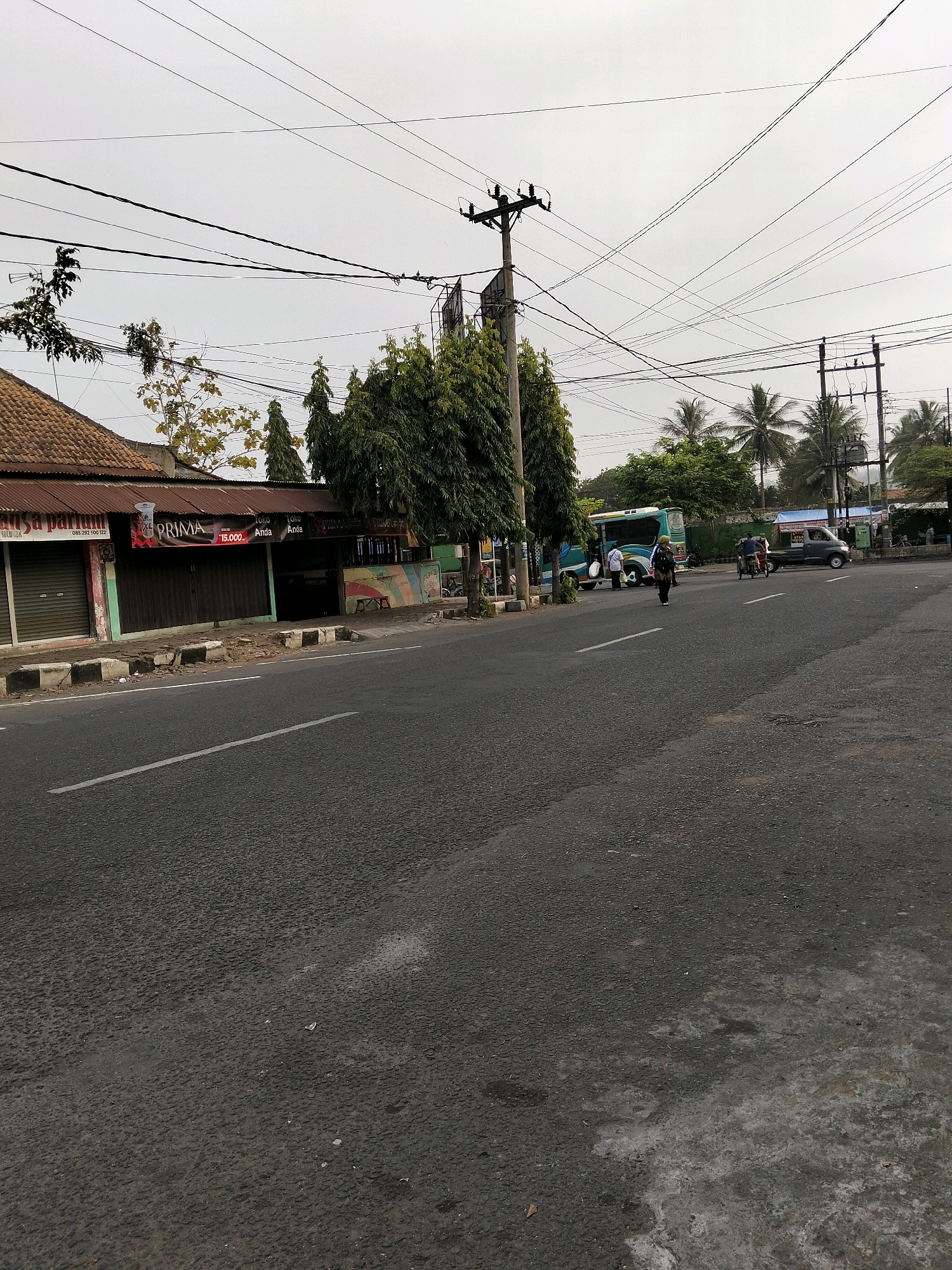 A wide shot shows a street scene in Purworejo, Indonesia, under an overcast sky. A two-lane asphalt road dominates the foreground, with a white dashed line marking the center. To the left, a row of shops with metal shutters line the sidewalk. One shop has a sign that reads "PRIMA" with a price of "15.000." To the right of the road, a blue and green bus is parked, and several people are visible. One person, dressed in dark clothing, walks across the street, while a few others stand near the bus. Further down the road, a dark gray pickup truck is also present. Telephone poles with numerous wires crisscross the sky above the scene. Palm trees and other foliage are interspersed throughout the background.