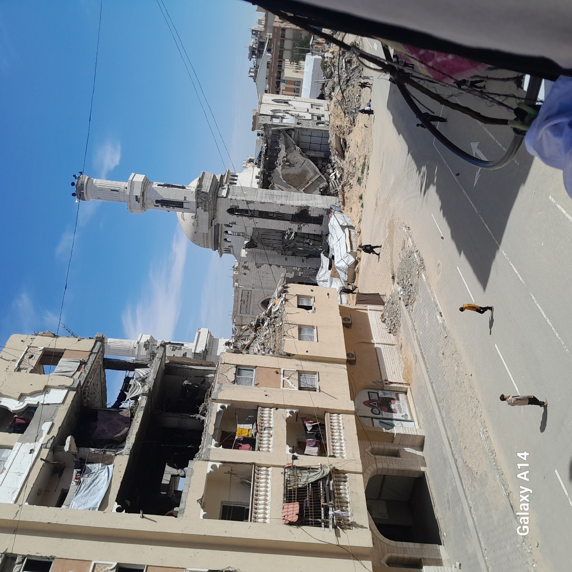 This image depicts a street scene in Khan Yunis, Palestinian Territory, showing extensive structural damage under a clear blue sky with sparse clouds.
In the foreground and left, a multi-story building exhibits severe damage, with large sections of its facade and internal structures missing, exposing interior rooms. Balconies on the remaining sections show laundry hanging, including white sheets and colored garments. The lower levels of this building feature arched entrances and some intact windows, with a partially visible mural on the ground floor.
To the right of the damaged building, further down the street, stands a severely damaged mosque. Its dome is partially collapsed, and one of its minarets is visibly compromised, with segments missing and structural elements exposed. Extensive rubble and debris surround the mosque, extending into what appears to be an open, sandy area. Several temporary shelters, constructed from white tarpaulins, are visible near the mosque's perimeter.
The street itself is paved with asphalt, marked with white lines, and appears relatively clean of large debris, though sandy patches are present on the shoulders. Scattered individuals are observed on the street and in the surrounding areas. Three individuals are walking on the paved street: one young male in a beige jacket and dark pants walks towards the left, another young male in a yellow shirt walks further down the street towards the right, and a third figure in dark clothing walks away from the camera in the mid-ground. Several other figures are visible near the mosque and in the debris-strewn areas, with two individuals appearing to interact or rest on the ground near a pile of rubble.
Power lines traverse the sky overhead. A partial view of a dark, curved object, possibly part of a fence or railing, is visible on the far right, partially obscuring the view. The watermark "Galaxy A14" is present in the lower left of the original image.