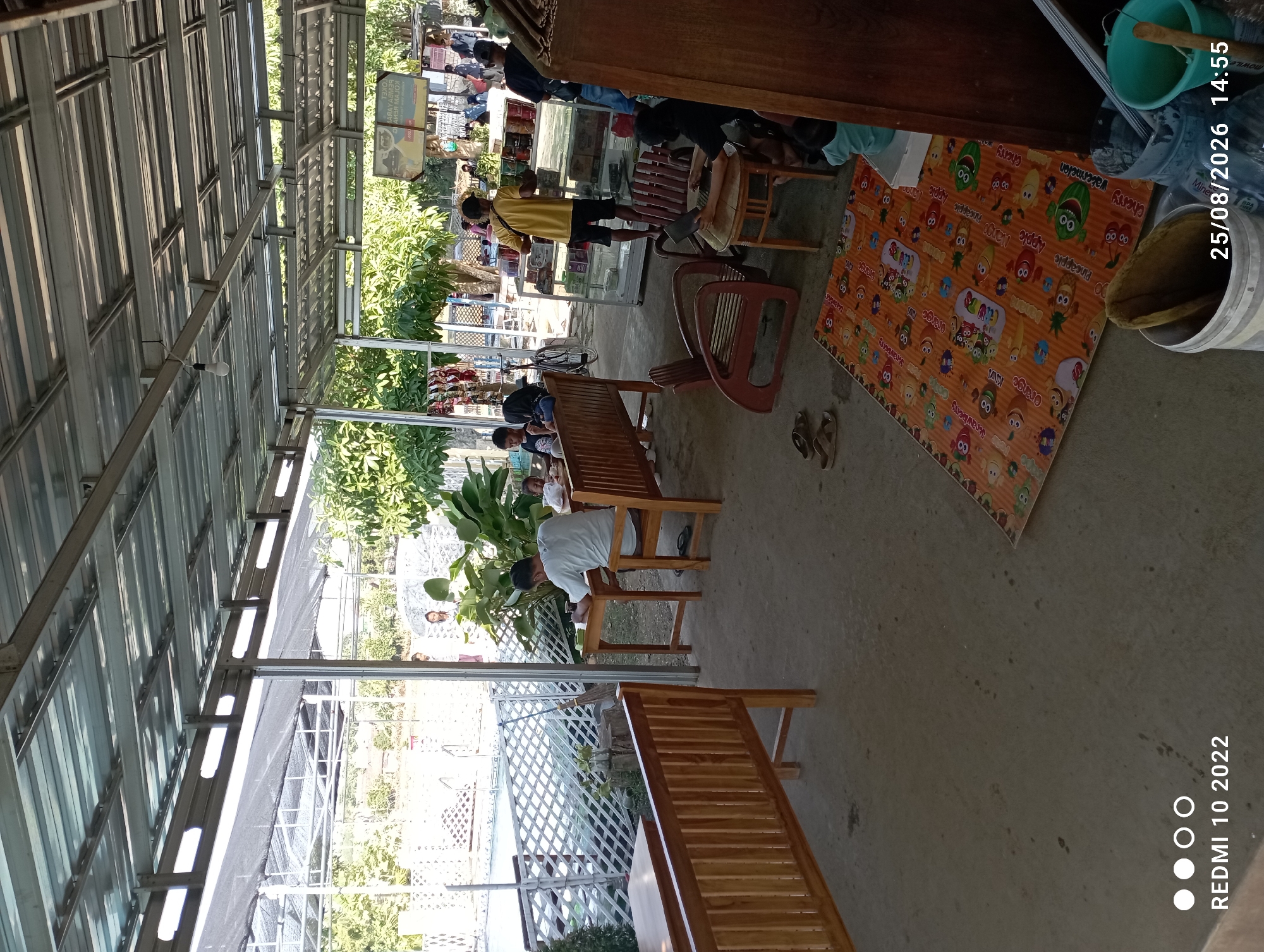 This image depicts an outdoor dining and recreational area, likely located in Tayu, Indonesia, as indicated by the timestamp and location in the lower left corner of the image. The scene is covered by a corrugated metal roof supported by metal beams, providing shade from the sun.
In the foreground, a long, wooden bench with a slatted backrest is positioned on a concrete floor. To the right, a person is seated at a small, round wooden table, appearing to use a laptop. Nearby, there is a wooden chair. Further to the right, a large wooden structure, possibly a cabinet or counter, is visible. A colorful mat with fruit illustrations is spread on the concrete floor. Two buckets, one white and one teal, are situated near the wooden structure, one containing what appears to be cleaning supplies.
In the midground, several wooden tables and benches are set up. One man in a white t-shirt is seated at a table, facing away from the camera, with a drink in front of him. Another table has a group of people, including children, seated around it. Behind these tables, a glass display case containing various items is visible, with a man in a yellow shirt standing in front of it.
In the background, a swimming pool is partially visible, with a white latticed fence separating it from the seating area. Lush green trees and foliage are present, suggesting a tropical or subtropical environment. Several people are seen walking around in the background, and some clothing items are hanging on display to the left of the glass case.
The overall impression is a casual, outdoor gathering place with amenities for dining and recreation. The lighting suggests it is daytime, with natural sunlight filtering through the roof and illuminating the scene.