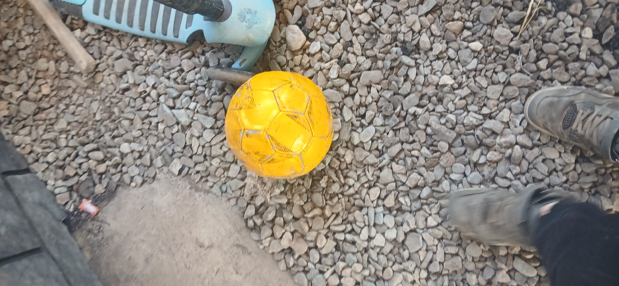 A high-angle outdoor shot shows a yellow soccer ball sitting on a gravel surface. To the right of the ball is a blue and black scooter. In the foreground, two feet are visible, one in a dark shoe and the other in a gray sneaker. The background features a concrete wall and more gravel. The scene is likely in Mihaylovka, Kyrgyzstan.