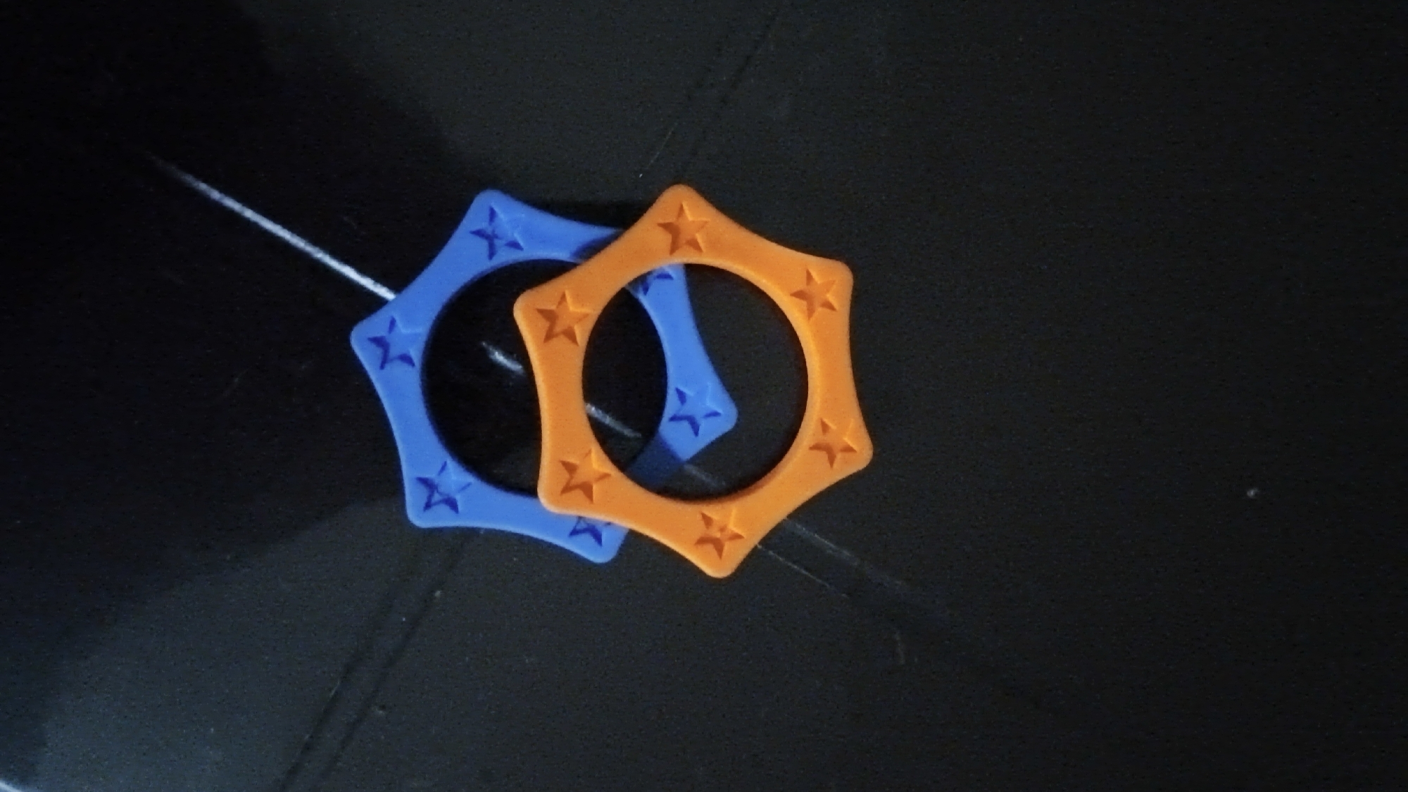 The image displays two star-shaped rings, one blue and one orange, placed on a dark, textured surface. The rings are positioned slightly overlapping each other. The surface appears to be a flat, dark material with subtle linear reflections, possibly indicating a polished or slightly uneven surface. The lighting in the image is dim, casting soft shadows and highlighting the textures of the objects. There are no people or any other objects visible in the frame. The scene is devoid of any context that would place it in a specific location within South Jakarta, Indonesia, or any other geographical area. The image solely focuses on the two star-shaped rings against a dark backdrop.