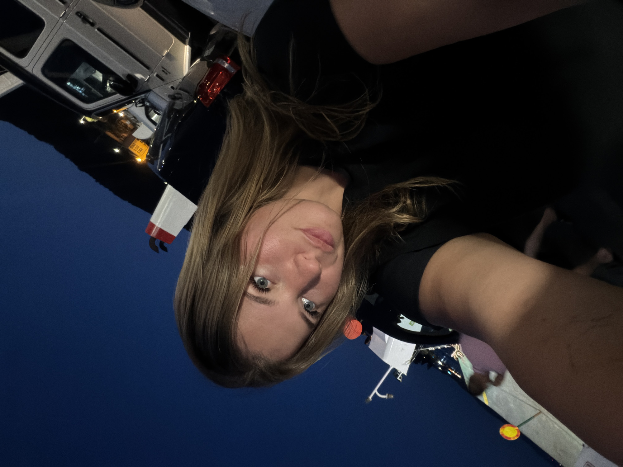 A young woman with blonde hair and blue eyes is taking a selfie outdoors at night, possibly on a ferry or similar transport with vehicles. The sky is dark blue, indicating evening or night. In the background, several vehicles, including a white SUV, are visible, some illuminated by streetlights or vehicle lights. The woman is wearing a black t-shirt and is positioned in the foreground, with her face turned towards the camera. There are some structures and lights in the mid-ground and background, suggesting an urban or port environment. The image is slightly tilted, giving a dynamic perspective. While the city of Đurići, Montenegro, is mentioned, the image itself does not contain explicit visual cues to confirm this specific location; the scene is general enough to be plausible in many port cities.
