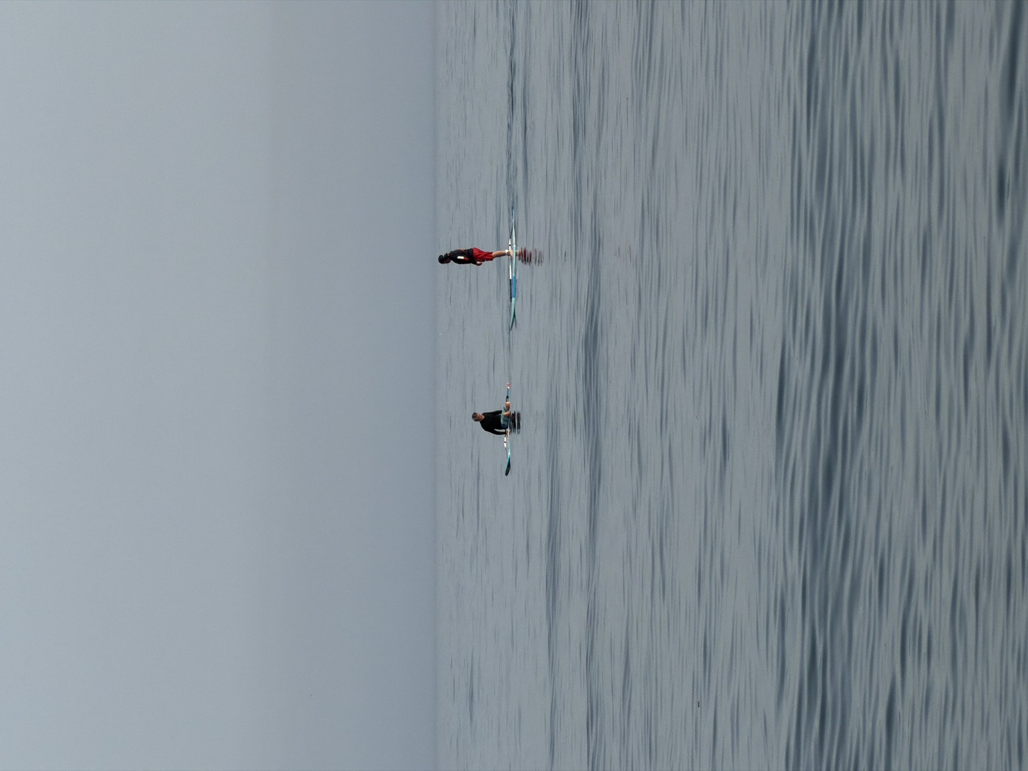 Two individuals are paddleboarding on a calm body of water under an overcast sky. The person on the left is seated on their paddleboard, wearing a black long-sleeved shirt and dark shorts. The person on the right is standing on their paddleboard, wearing a black long-sleeved shirt and red shorts. Both paddleboards are oriented horizontally. The water is a muted blue-grey with gentle ripples. The horizon line is indistinct due to the fog or haze. The scene appears to be a coastal area, possibly the Mediterranean Sea off the coast of Estepona, Spain.