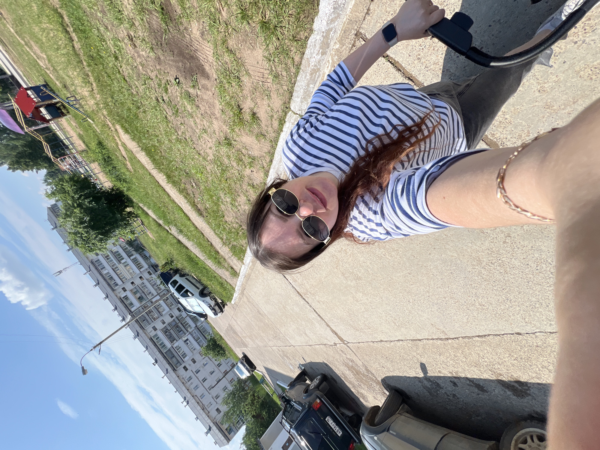 This media file is a slightly tilted, first-person perspective selfie taken outdoors on a sunny day in Noviy, Russia. The subject is a young woman with long, dark brown hair, wearing a blue and white striped t-shirt and sunglasses. She is holding onto the handlebars of a stroller or bicycle.
The scene is a paved walkway next to a grassy area with a playground visible in the background. To the left of the walkway, there is a multi-story apartment building with numerous windows, characteristic of a residential area. Several cars are parked along the side of the building. The sky is blue with scattered white clouds, suggesting it's daytime. The sunlight creates shadows on the ground, indicating a bright and likely warm day. The overall mood appears casual and relaxed, consistent with a person enjoying an outdoor activity.
There is a license plate on one of the parked cars with the visible text "A816YY 16". The watch on the woman's left wrist appears to be a smartwatch.