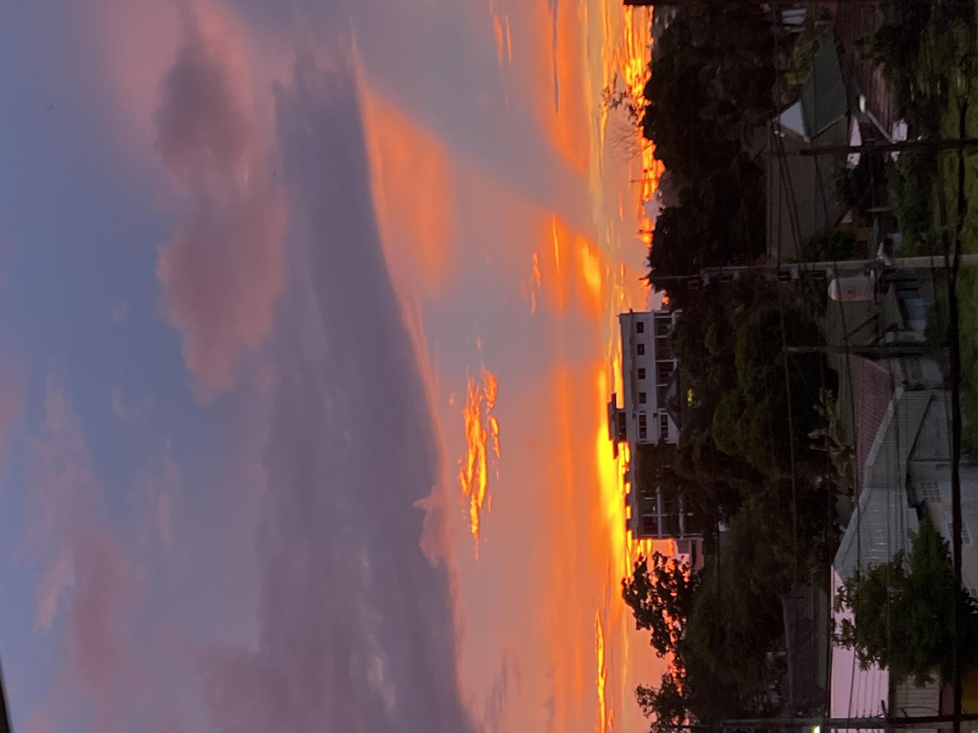 This is a vertical, eye-level, medium shot of a vibrant sunset over a city. The sky dominates the frame, showcasing a dramatic gradient of colors from deep blue and purple at the top to fiery orange and yellow near the horizon. Wispy clouds are illuminated by the setting sun, creating streaks of bright light and casting dramatic shadows across the sky.
Below the sky, a multi-story building with a white facade and visible windows stands prominently. It is surrounded by dense, dark green trees. In the foreground, several rooftops of houses are visible, some with peaked roofs and others with flat surfaces. The scene appears to be set in a residential area with overhead power lines crisscrossing the lower portion of the image. The overall impression is one of a tranquil, urban evening scene bathed in the warm glow of the setting sun.