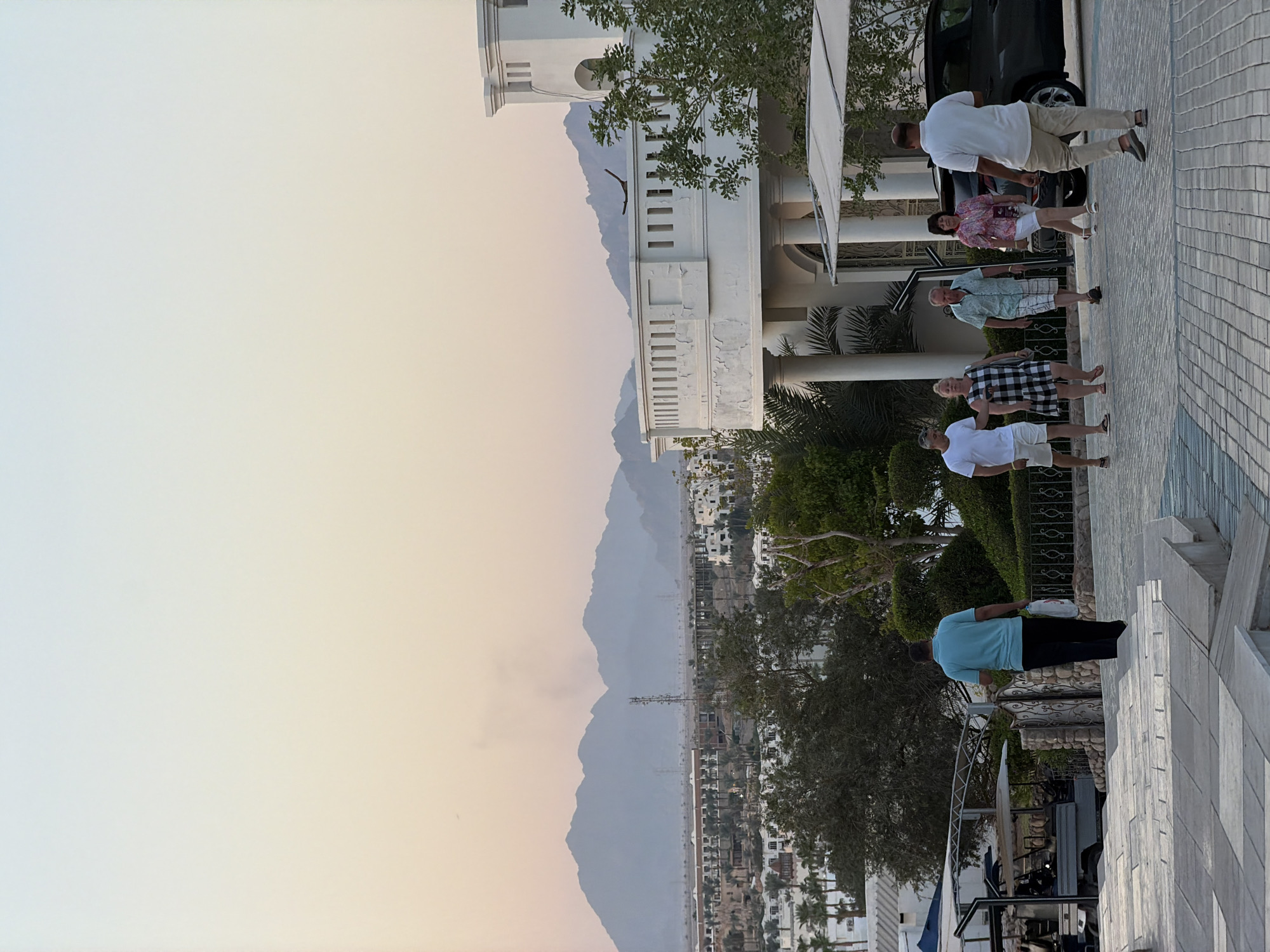 The image depicts a group of people walking along a paved pathway in Sharm El Sheikh, Egypt, with a resort complex and mountains in the background. In the foreground, a man in a turquoise shirt and black pants walks away from the camera, carrying two white plastic bags. To his right, five individuals walk towards the camera. From left to right, they are a man in a white t-shirt and white shorts, a woman in a black and white checkered dress, a man in a light green patterned shirt and white shorts, a woman in a pink floral shirt and white shorts, and a man in a white polo shirt and khaki pants. Behind the walking group, a large white building with columns and a balcony is visible, part of a resort complex. Palm trees and other lush greenery are scattered throughout the scene. In the distance, a range of hazy mountains forms the backdrop under a pale, gradient sky that transitions from light blue to peach. The overall scene suggests a warm, possibly late afternoon or early evening setting at a tourist destination.