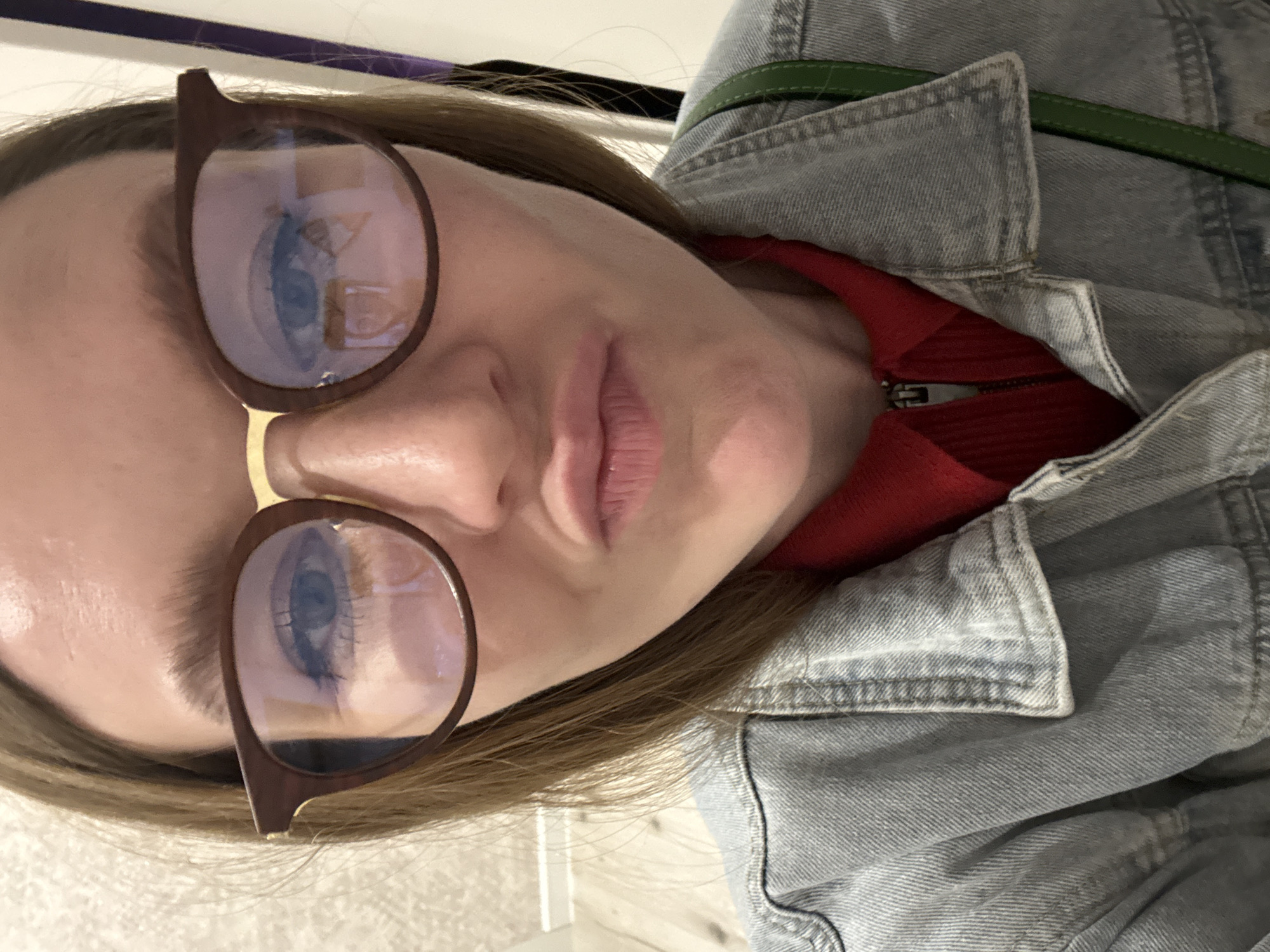 A close-up selfie shows a young woman with brown hair, wearing large, round, brown-rimmed glasses with gold accents. She has fair skin, visible eyelashes, and is making a pouty facial expression. She is wearing a red collared shirt with a zipper closure and a light blue denim jacket. A green strap from a bag is visible over her right shoulder. The background appears to be an indoor setting with a textured wall on the left and a vertical element on the right that could be part of a doorway or a poster. There are reflections in her glasses that suggest the presence of a light source and possibly other objects in the room, such as a window or a screen. There are no identifiable landmarks or contextual clues to suggest the location is Izhevsk, Russia.