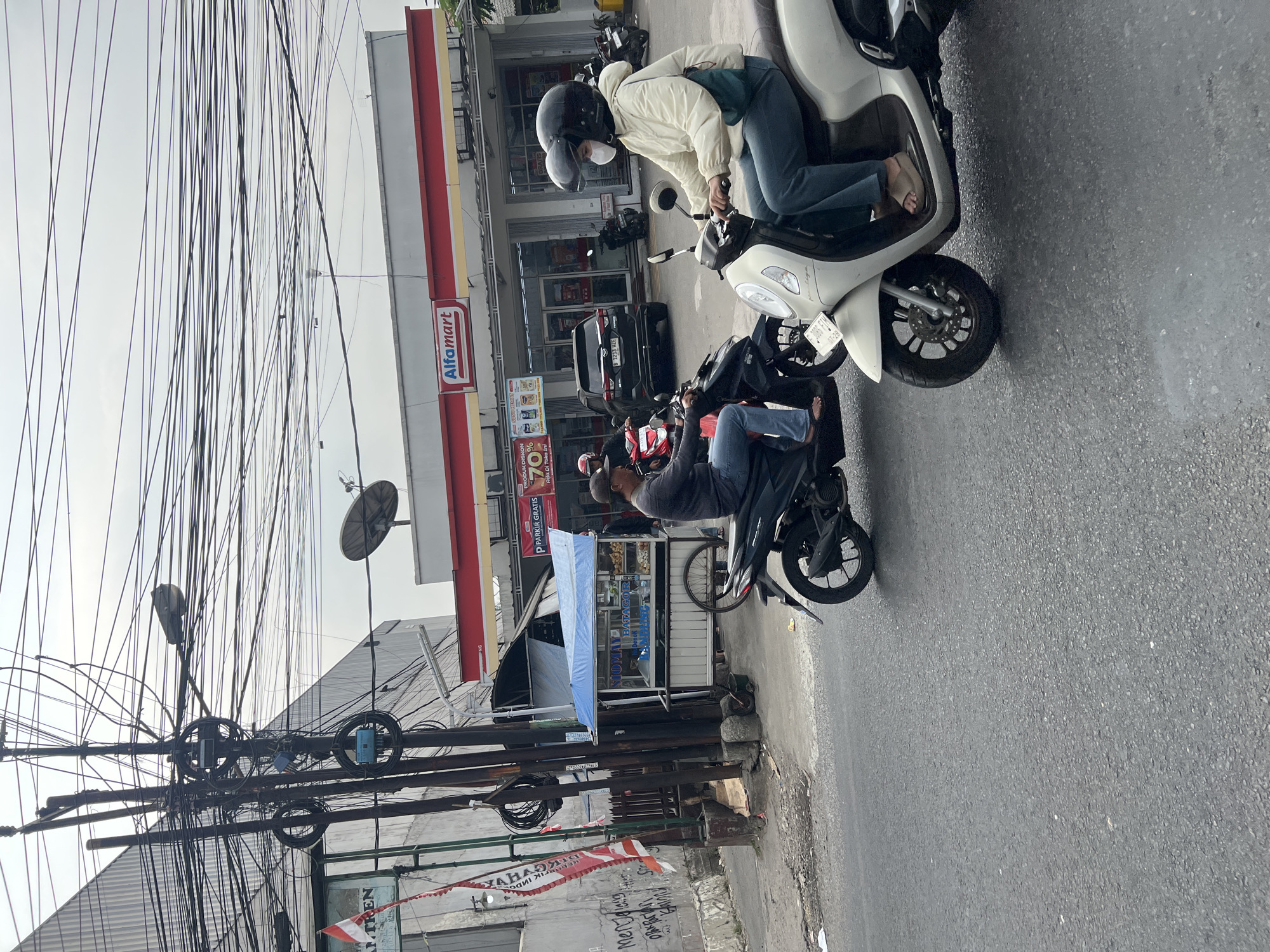 The image depicts a street scene in Citeureup, Indonesia, with a prominent Alfamart convenience store in the background. Multiple motorcycles are present, with two individuals riding in the foreground. One motorcycle is white, ridden by a person wearing a white jacket, blue pants, a helmet, and a mask. They are wearing sandals. Another motorcycle, black, is ridden by a person in a dark jacket and blue jeans, also wearing a helmet.
In front of the Alfamart, a black car is parked. A food stall with a blue awning is set up on the sidewalk. The environment is characterized by a sky filled with a dense network of electrical wires, typical of urban Indonesian settings. The road is paved and appears wet, suggesting recent rain or a damp surface. Graffiti is visible on a wall to the left of the scene. A satellite dish is mounted on the roof of a building.