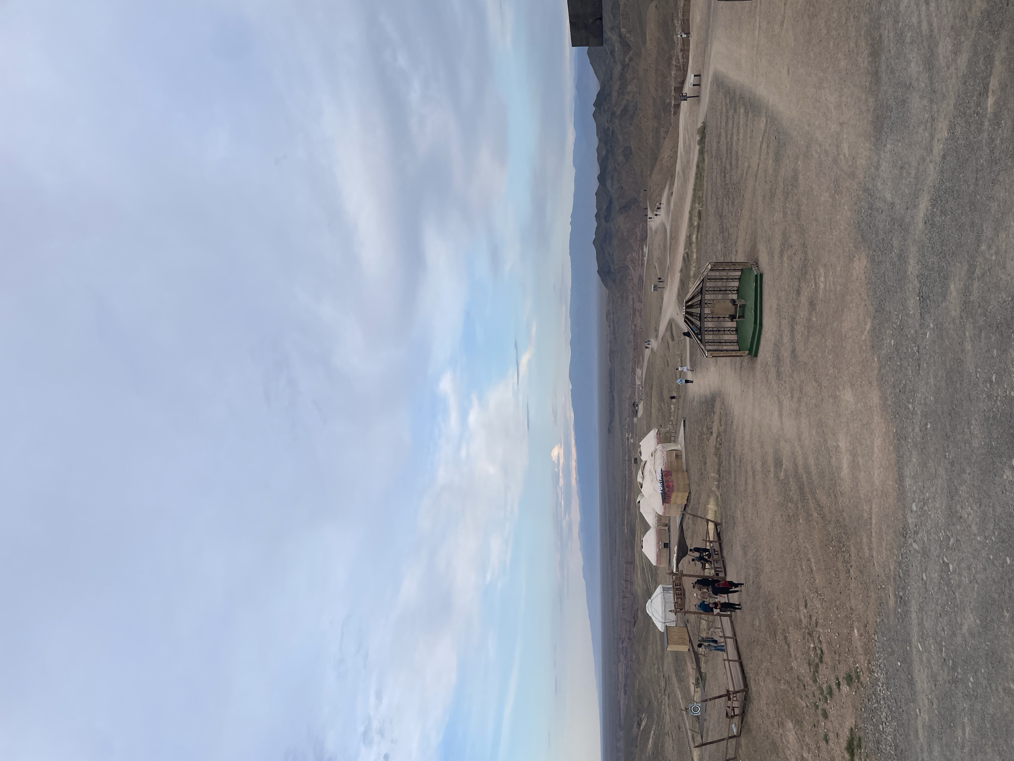 The image depicts a vast, arid landscape in the Charyn National Park, near Tamerlik, Kazakhstan, under a cloudy sky. In the foreground, a dirt path winds through the dry, rocky terrain. Several yurts, traditional nomadic dwellings, are clustered together, some with visible signage. A group of people are gathered near a wooden structure with a target, suggesting an activity area. Further in the distance, more yurts and scattered individuals can be seen along winding paths. The background is dominated by rolling hills and distant mountains, characteristic of the Charyn Canyon region. A decorative, open-sided structure with a chair inside sits on a green mat in the middle ground, adding a unique element to the scene. The overall impression is one of a remote, tourist-oriented location in a rugged natural environment.