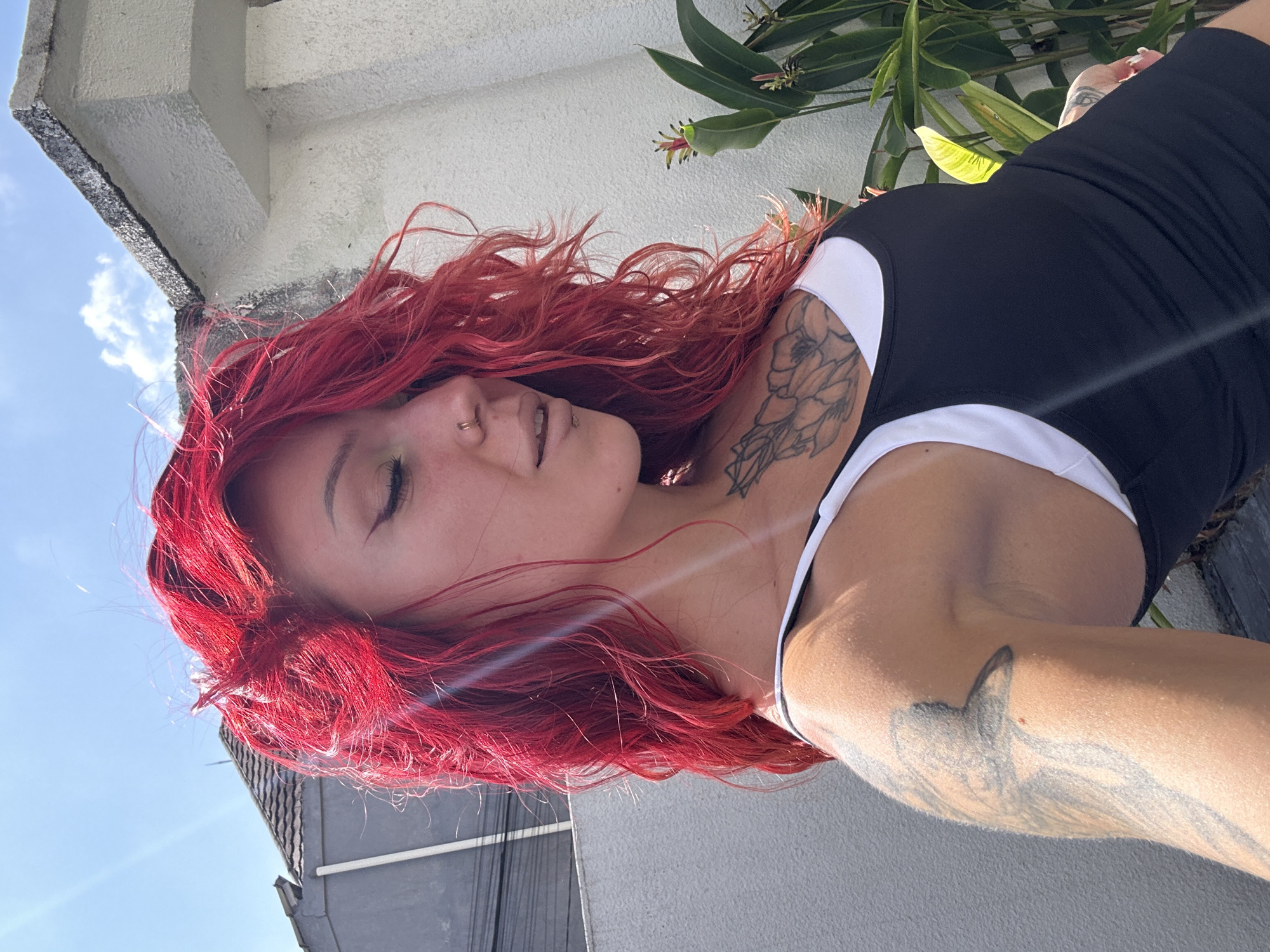 A woman with bright red, wavy hair is captured in a close-up, profile shot outdoors. She is wearing a black tank top over a white sports bra. Tattoos are visible on her chest and arm. Her head is tilted downwards and to the side, with her eyes closed. The background features a light-colored wall, a grey building with a pitched roof, and some greenery, including a plant with red and yellow flowers. The scene appears to be in an urban or suburban setting, possibly a residential area in North Kuta, Indonesia, indicated by the architectural style and vegetation. The lighting suggests it is daytime, with sunlight casting shadows and highlights.