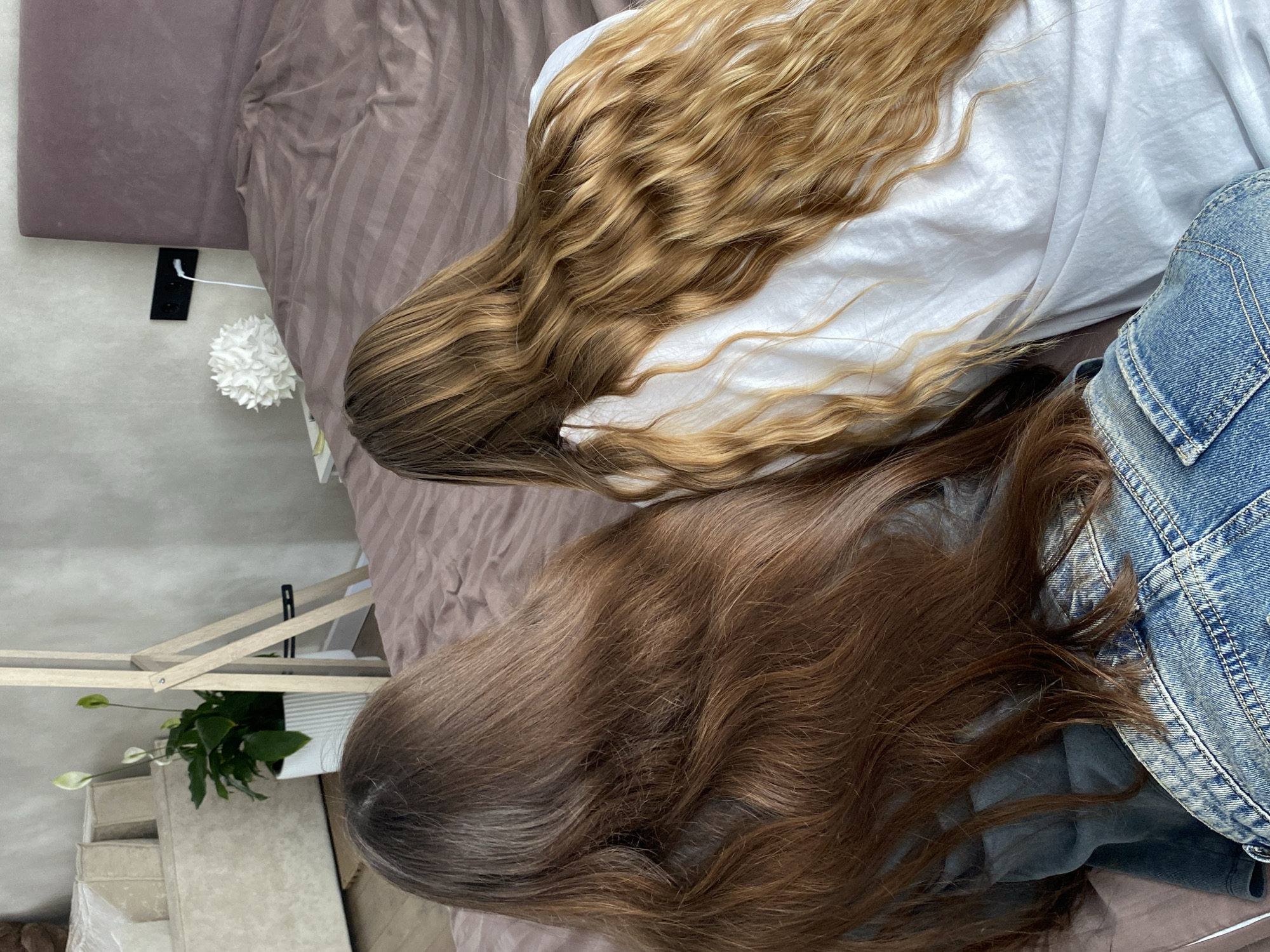 A top-down view shows two individuals lying on a bed. The individual to the left has long, brown hair, while the other individual has long, blonde hair and is wearing a white shirt and denim jacket. They are lying on a bed covered in brown sheets. A white flower decoration and a floor lamp with a plant are visible in the lower left corner. The location cannot be determined solely from the image.
