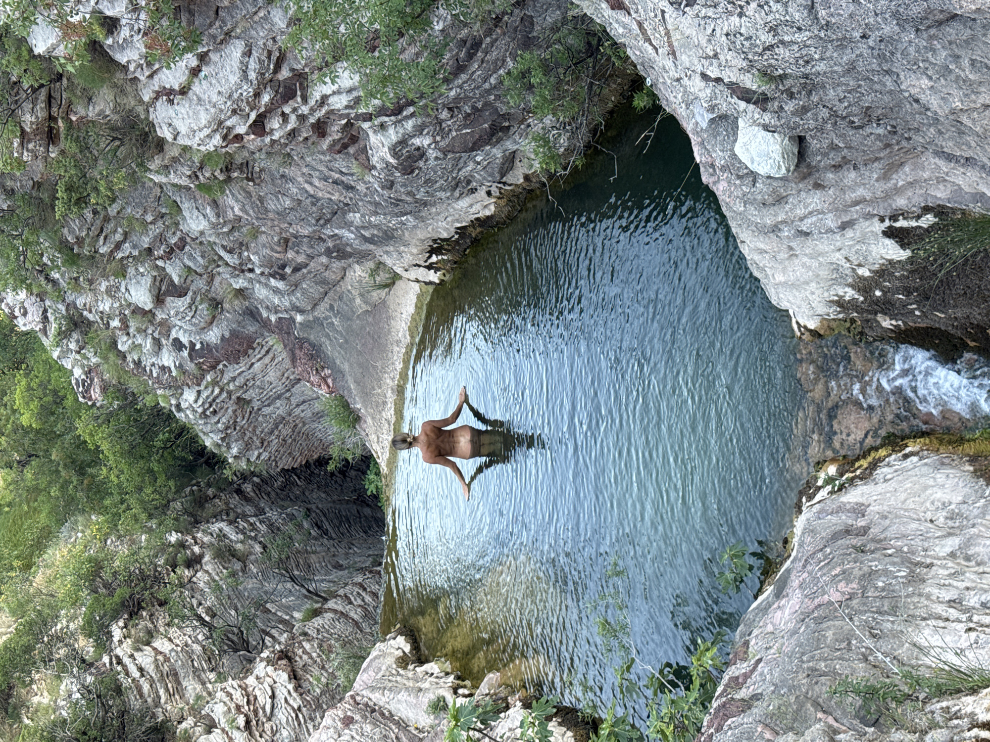 A person stands in the middle of a natural pool of water surrounded by rocky cliffs covered in vegetation. The person is facing away from the camera, with their arms outstretched and resting on the water's surface. The water is clear and reflects the surrounding environment. The cliffs are made of stratified rock with varying colors and textures, and small trees and bushes grow on their surfaces. A small waterfall is visible at the bottom of the pool, cascading over rocks. The scene appears to be a secluded natural oasis.