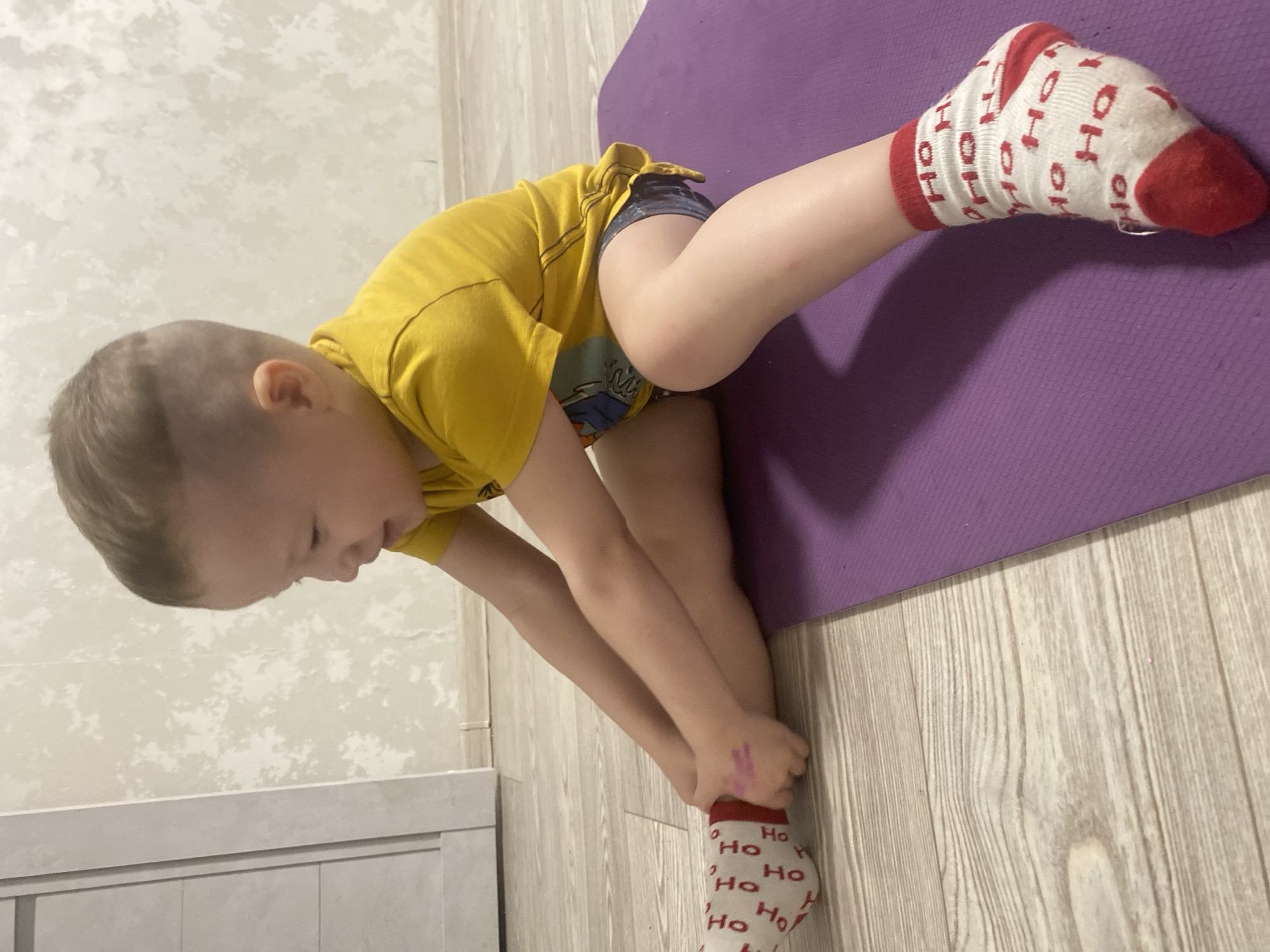 A child in a yellow shirt and denim shorts is stretching on a purple yoga mat indoors. The child is bending forward, holding the foot of their leg that is on the floor. Both feet are wearing white socks with red trim and "Ho" printed on them. The floor is light-colored wood-grain laminate. A white cabinet is visible in the background. The image does not suggest a location in the city of Sochi, Russia.