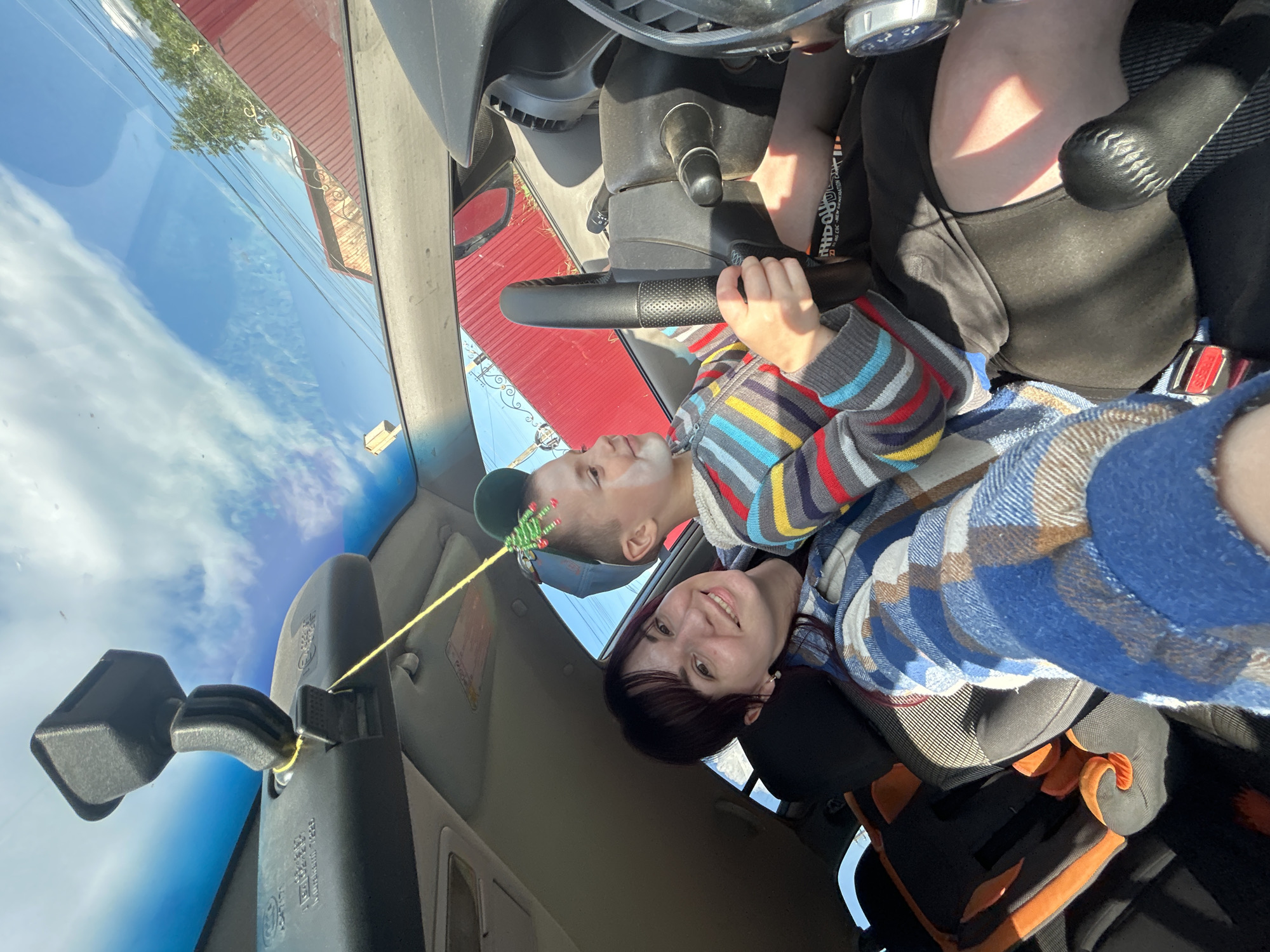 A young woman with dark hair and a blue plaid shirt smiles as she sits in the passenger seat of a car. Next to her, a young boy with a cap and face paint grips the steering wheel, appearing to mimic driving. A yellow string hangs from the rearview mirror with a small green and red decorative item attached. The car's interior is visible, including a child car seat in the back. Outside the car, a red fence and trees are visible under a blue sky with white clouds. The scene appears to be a sunny day.