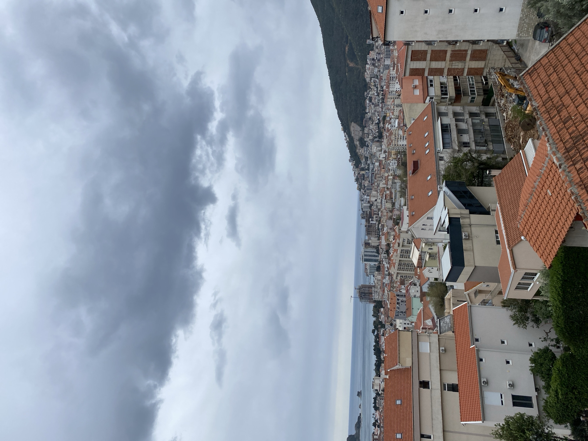 The image depicts a cityscape under a cloudy sky, likely taken from an elevated vantage point. The location is Budva, Montenegro, as evidenced by the architecture.
The foreground features buildings with terracotta-colored roofs and light-colored facades. In the middle ground, there's a dense arrangement of structures, including apartments, houses, and hotels. The sea stretches along the horizon, dividing the urban landscape from the sky. A mountain runs parallel with the city and sea. The sky is heavily overcast with thick, gray clouds dominating the frame. An excavator can be seen within the city.