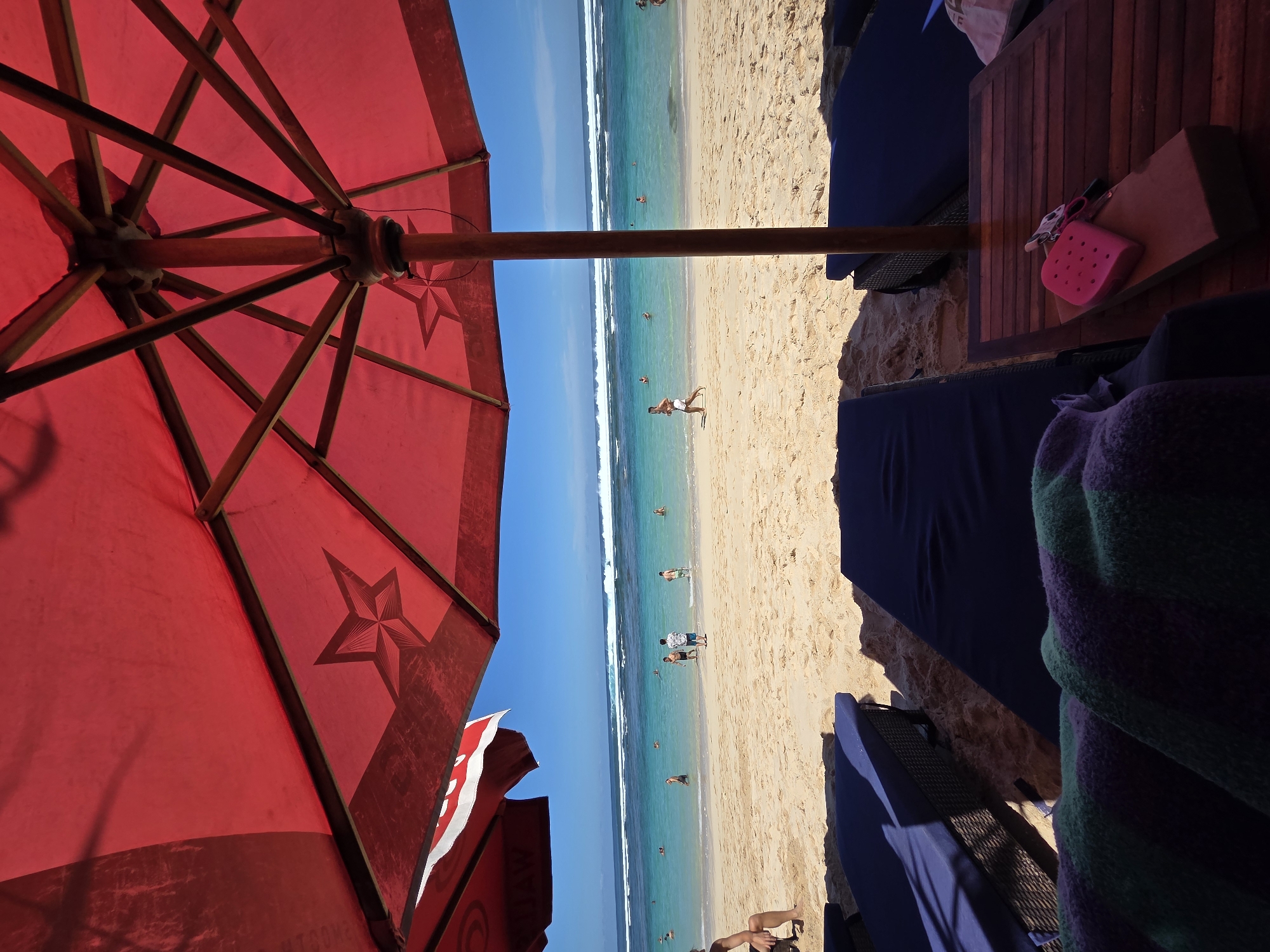 The scene depicts a sunny beach in Kuta Selatan, Indonesia, viewed from beneath a large red beach umbrella. The umbrella has a wooden frame and features a star design. The beach is crowded with people swimming and walking along the shore. The water is clear and turquoise. To the right of the frame, there is a wooden table with a pink sponge on it, beach chairs, and a folded towel. The image appears to be taken from a first-person perspective, sitting under the umbrella.