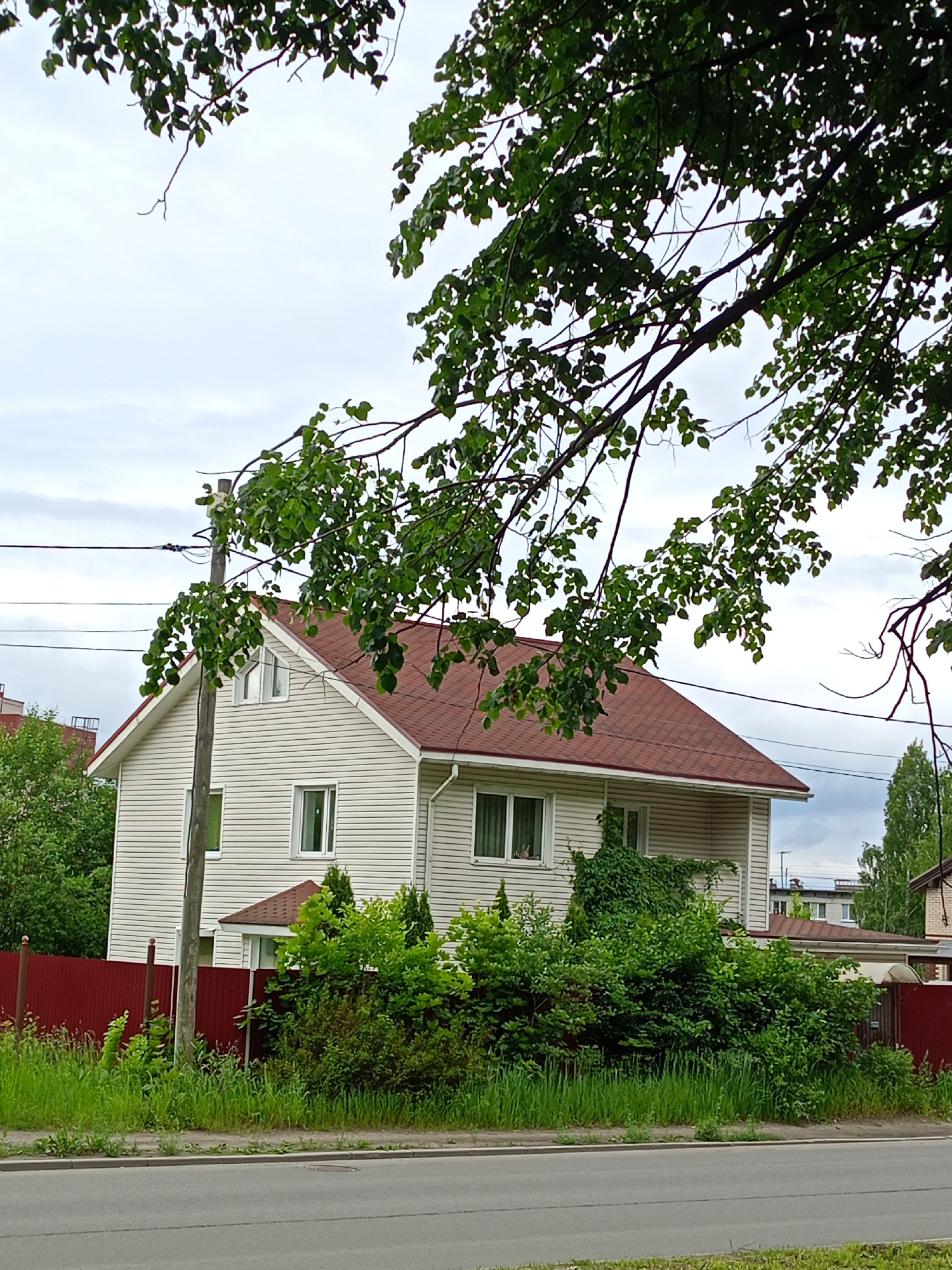 A two-story, cream-colored house with a red roof sits behind a red fence and dense green foliage. The house has white-trimmed windows and what appears to be a vinyl siding exterior. A large tree with green leaves partially obscures the top of the house and the sky, which is overcast. A wooden utility pole stands in front of the house. A paved road with a grassy shoulder runs along the bottom of the image. The scene appears to be in a suburban or semi-rural setting.