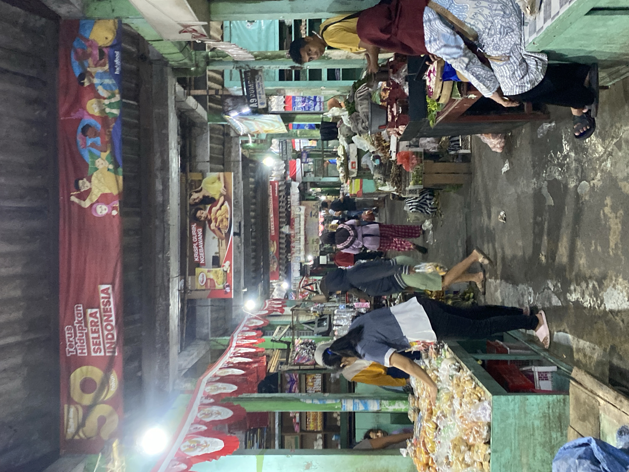 The image depicts a bustling market scene in Ambarketawang, Indonesia. A long, covered walkway lined with stalls showcases a variety of goods, primarily food items displayed on long, green counters. Numerous individuals, including vendors and shoppers, are present, engaged in transactions and navigating the crowded space. Some vendors are seen attending to their merchandise, while others interact with customers. The environment appears somewhat weathered, with visible signs of wear and tear on the structures and a slightly damp floor, suggesting recent rain or regular cleaning. Overhead, banners and decorations are hung, contributing to the vibrant, communal atmosphere of the market.