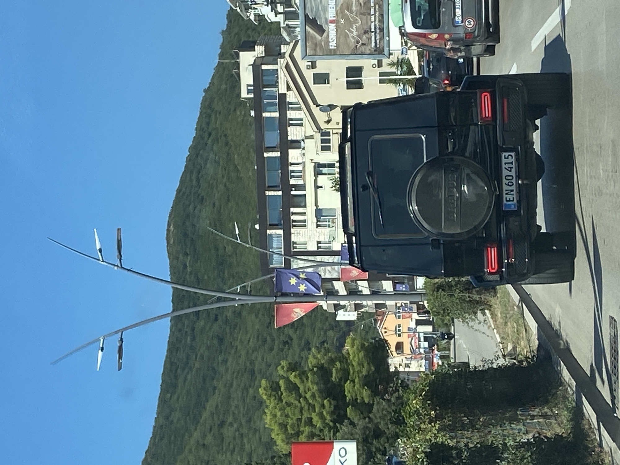The image shows a street scene in Budva, Montenegro. The foreground features the rear of a black Mercedes-Benz G-Class SUV with a spare tire mounted on the back. The license plate reads "EN 60 415". In front of the SUV are two lanes of traffic, with another vehicle visible in the right lane. The background consists of buildings, a lush green hillside, and streetlights with decorative banners. A billboard with "FASHION FRIEND" is visible on the right side. The sky is clear and blue.