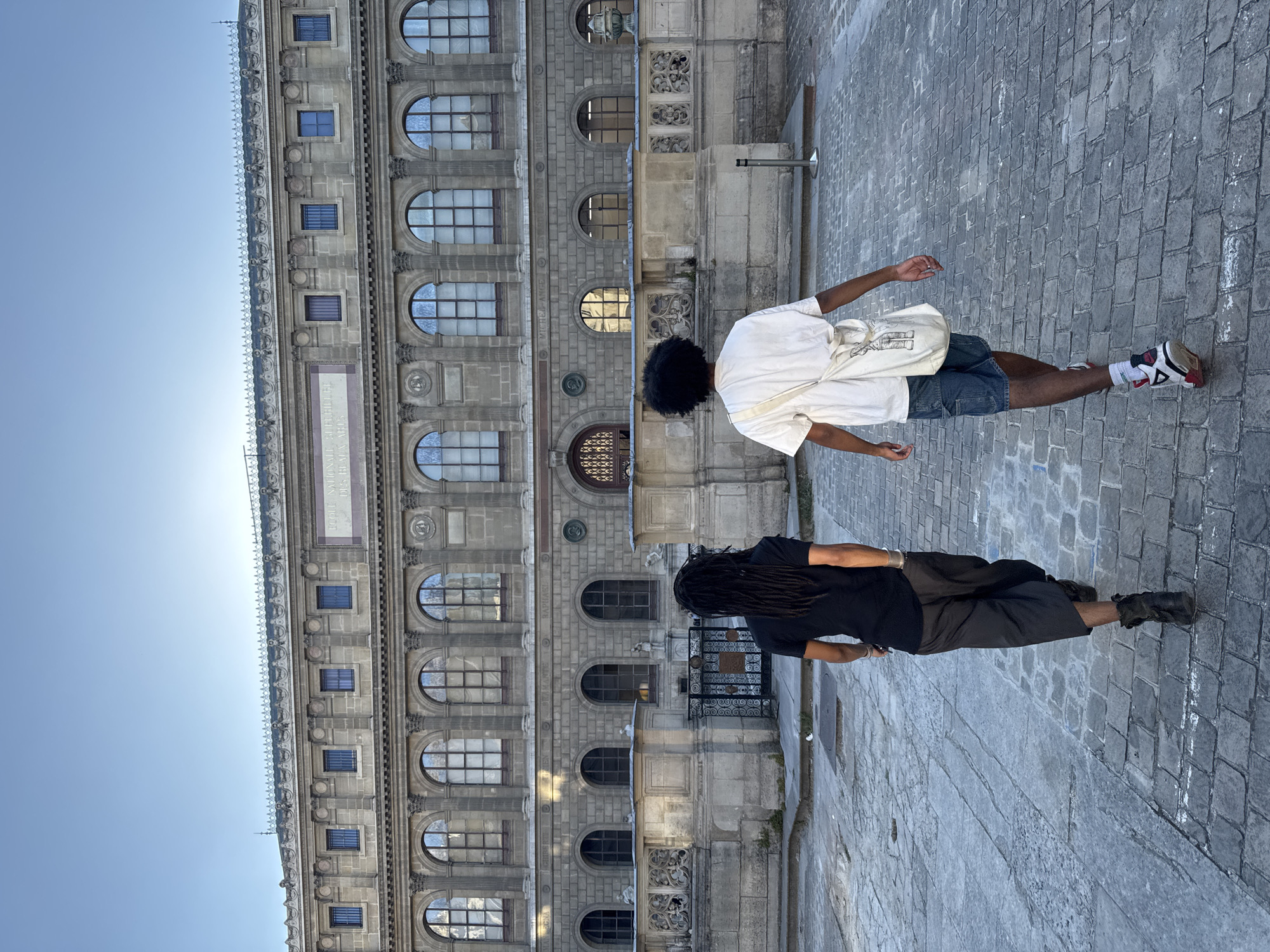 Two people stand on a stone-paved street in front of a large stone building. The person on the left wears a black t-shirt, dark-colored shorts, and black shoes. The person on the right wears a white t-shirt, blue denim shorts, a white bag, and red/white sneakers. The background building features rows of arched windows and ornate stonework. Based on its architectural style and location, the building is the Bibliothèque Nationale de France (National Library of France) in Paris.