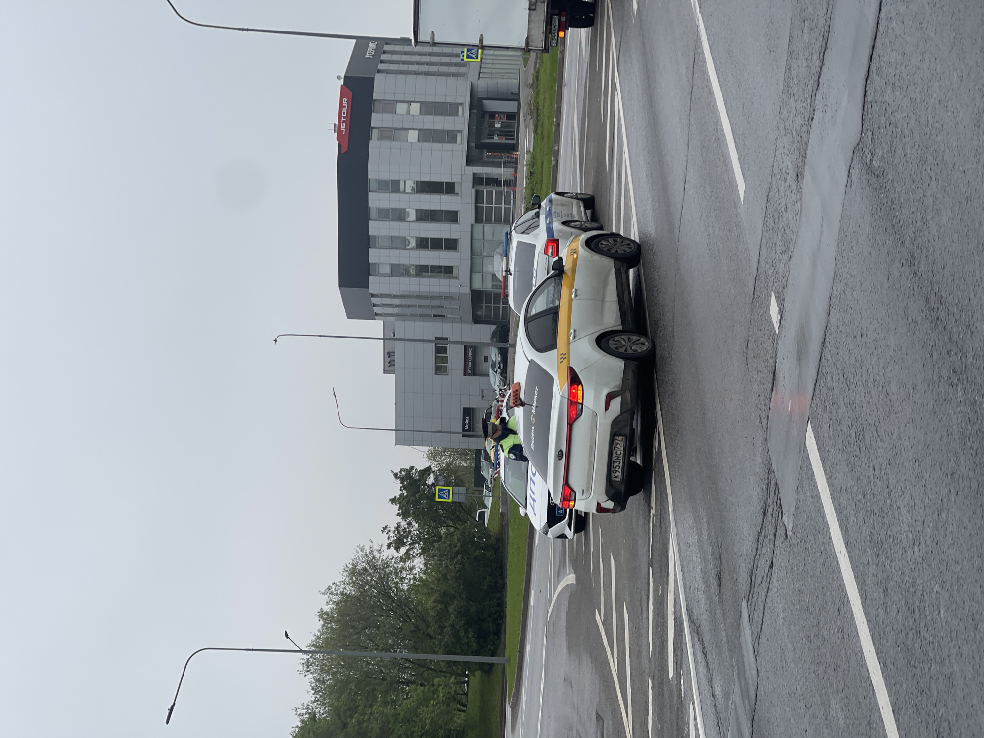 The image depicts a street scene in Khimki, Russia, on an overcast day. A white taxi with a yellow stripe, identified by its license plate number K953HC797, is stopped on a multi-lane road. Behind the taxi is a police car with a flashing blue light on top. An officer in a green vest is standing between the two vehicles. The building across the street has the logo "JETOUR" and "СЕРВИС" on it.