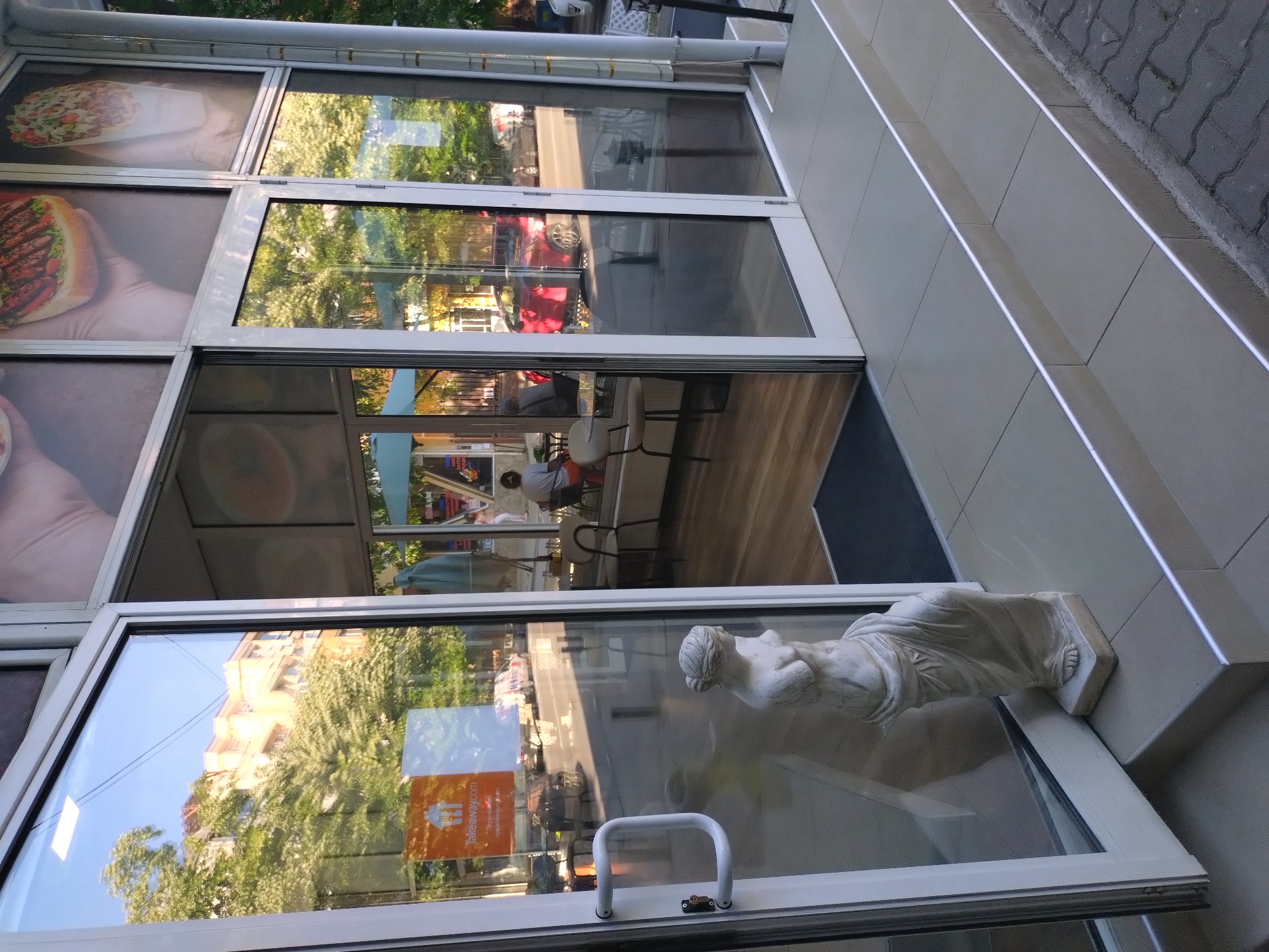 This image captures the entrance to a commercial establishment in Stara Zagora, Bulgaria, characterized by a glass facade and three tiled steps leading up from the sidewalk. The entrance consists of white-framed glass doors, the left door being slightly ajar and propped open by a white classical-style statue of a draped female figure, lacking a head and arms, standing on the bottom step. The doors feature white horizontal handles. Above the doors, large glass panels display advertisements for food, including a close-up of a burger held by hands and a cone of mixed ingredients. A "Takeaway.com" sticker with additional Cyrillic text is affixed to the lower left glass door.
Reflections on the glass panels reveal the surrounding urban environment. The upper left glass reflects a multi-story building with a light facade, dense green trees, and a clear blue sky with overhead wires. The lower glass panels reflect a street with vehicular traffic, including a prominent red car, and other parked vehicles.
Through the clear glass, the interior of the establishment is visible, showing a light-colored wooden floor. Tables and chairs are arranged inside, and at least two individuals are present: one person in a gray shirt and orange trousers is seated at a table on the left, facing away, and another person is seated further inside. The lighting suggests it is daytime. The steps leading to the entrance are constructed of light-colored tiles, each edged with a thin, reflective metallic strip. The ground beyond the steps is paved with dark grey interlocking bricks.