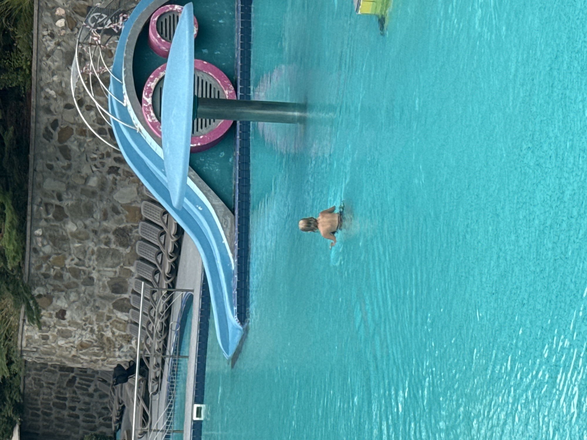 The image shows a swimming pool with a blue water slide. A young child with blonde hair is swimming in the pool, facing away from the camera. In the background, there are lounge chairs and a stone wall. A cylindrical structure with a disc-shaped top is submerged in the pool near the slide. The environment appears to be an outdoor recreational area.