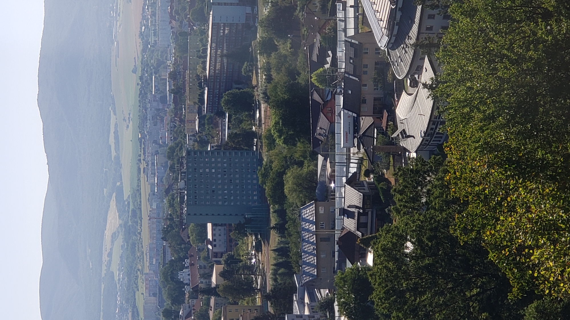The image is a high-angle, eye-level shot of a cityscape with mountains in the background. The foreground is dominated by lush green trees and foliage, with hints of residential buildings peeking through. In the middle ground, there are several multi-story buildings, including a prominent light blue high-rise, and various houses with pitched roofs. Further in the distance, a vast urban sprawl stretches towards rolling, hazy green hills and mountains. The sky is clear and pale blue. A truck with "PEDMONT" written on its side is visible in the middle ground, suggesting ongoing transportation or logistics activity. The overall impression is of a densely populated area nestled within a natural landscape. Based on the architectural style and the general environment, this scene is consistent with views from the Sídlisko III neighborhood of Prešov, Slovakia, looking towards the surrounding hills and mountains.