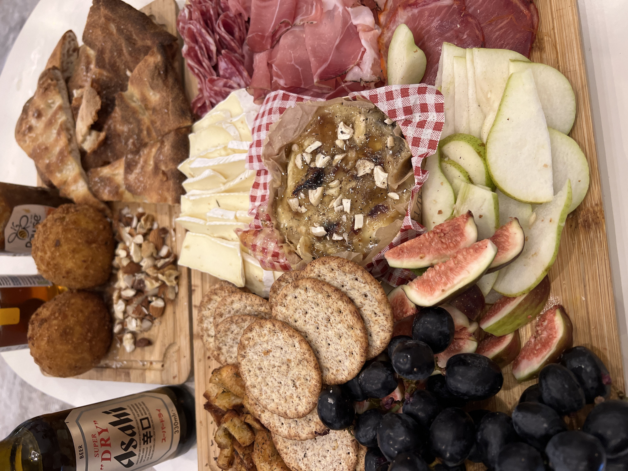 The image shows a charcuterie board filled with a variety of food items. The board is divided into sections, each containing different types of food. There is a section with bread, a section with cured meats (like salami and prosciutto), a section with cheese, a section with crackers, a section with nuts, a section with fried appetizers, and a section with fruit (grapes, pears, and figs). A bottle of "Asahi Super Dry" beer is also visible on the left side of the frame. The board is arranged on a white surface, likely a table or countertop.