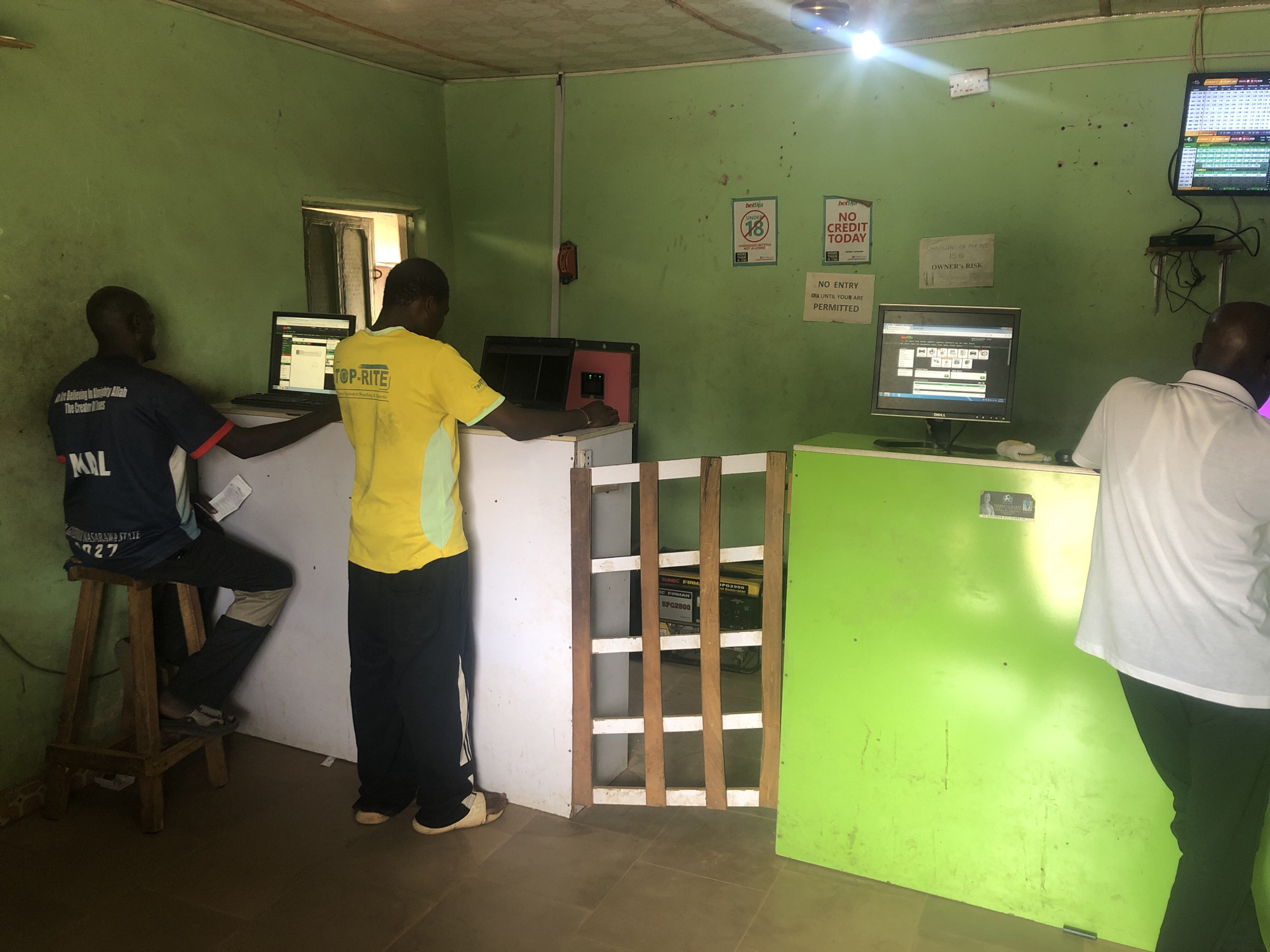 The image captures an indoor scene, likely a betting shop or internet cafe, in Akwanga, Nigeria. The room has light green walls, a light-colored ceiling, and tiled flooring. The overall impression is utilitarian and functional, with some wear and tear visible on the walls.
Three men are present. On the left, a man wearing a dark blue and white t-shirt with "MIL" and "NASARAWA STATE" text is seated on a wooden stool, looking at a laptop screen. He appears to be engaged with the computer. In the center, another man, wearing a yellow "TOP-RITE" t-shirt and dark trousers, stands behind a counter, also facing a computer screen. To the right, a third man in a white t-shirt and dark trousers stands with his back mostly to the camera, looking at a desktop monitor.
Multiple computer screens are visible, displaying interfaces that appear to be for online betting or gaming. One desktop monitor on the right shows a grid of data, possibly sports fixtures or results. Another monitor displays a website with icons, potentially a betting platform.
Notable objects include a wooden stool, counters, a wooden barrier-like structure dividing the space, and a generator (SUMEC FIRMAN SPG2800) visible behind the barrier. Bright overhead lights illuminate the room.
Several signs are affixed to the green wall:
* A "bet9ja" sign indicating "UNDER 18 SYMBOL BETTING IS NOT ALLOWED."
* Another "bet9ja" sign stating "NO CREDIT TODAY."
* A sign warning, "CHARGING OF PHONE IS @ OWNER'S RISK."
* A sign reading, "NO ENTRY ONLY UNTIL YOUR ARE PERMITTED."
* Text on the blue shirt reads, "e Believing in almighty Allah The Creators MIL NASARAWA STATE 27."
* Text on the yellow shirt reads, "TOP-RITE."
The activity appears to be focused on sports betting or similar online activities. The time of day is not explicitly clear, but the artificial lighting suggests it could be any time the establishment is open.