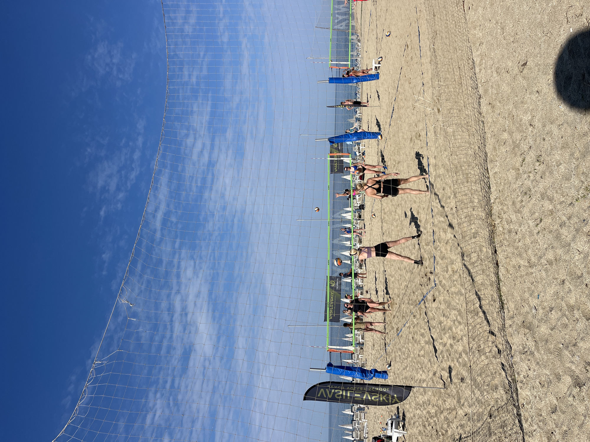 The image depicts a beach volleyball game being played on a sandy beach under a clear blue sky in Alanya, Turkey. The volleyball net is prominently visible, dividing the court where players are actively engaged in the game. Several individuals are captured in various stages of play, some lunging to hit the ball. Beach chairs and umbrellas are set up in the background, suggesting a recreational area. A banner with the text "VASILEVSKIY BEACH VOLLEYBALL SCHOOL" is visible. The sand appears dry, and the sunlight casts shadows of the players and the net.