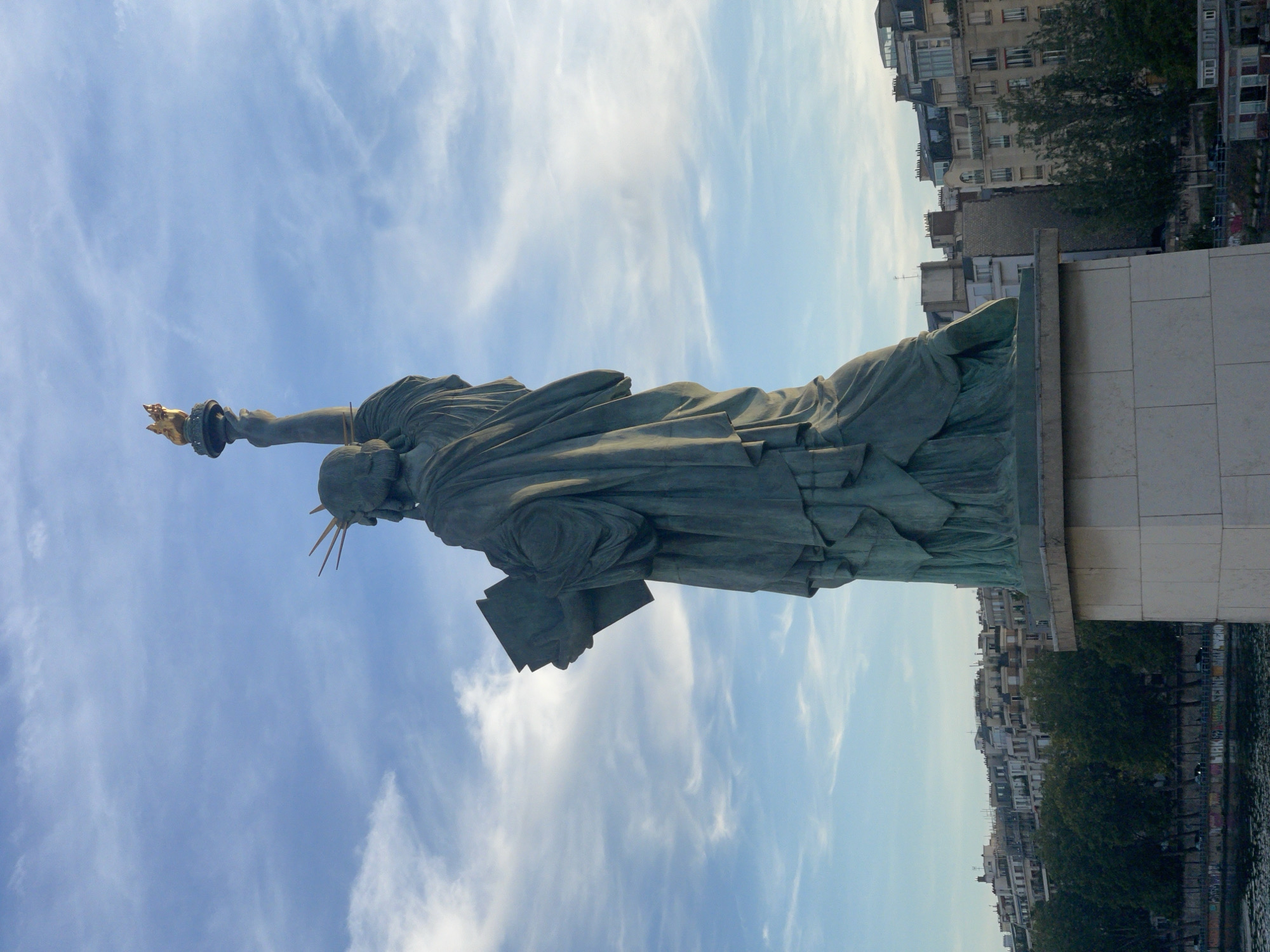 The image shows a statue of Liberty, viewed from the back, holding a torch and a book. The statue is made of bronze and is positioned on a stone pedestal. In the background, there is a city skyline with buildings and trees, and a body of water in the foreground. The sky is blue with some clouds. This statue is located on the Île aux Cygnes in Paris, France.