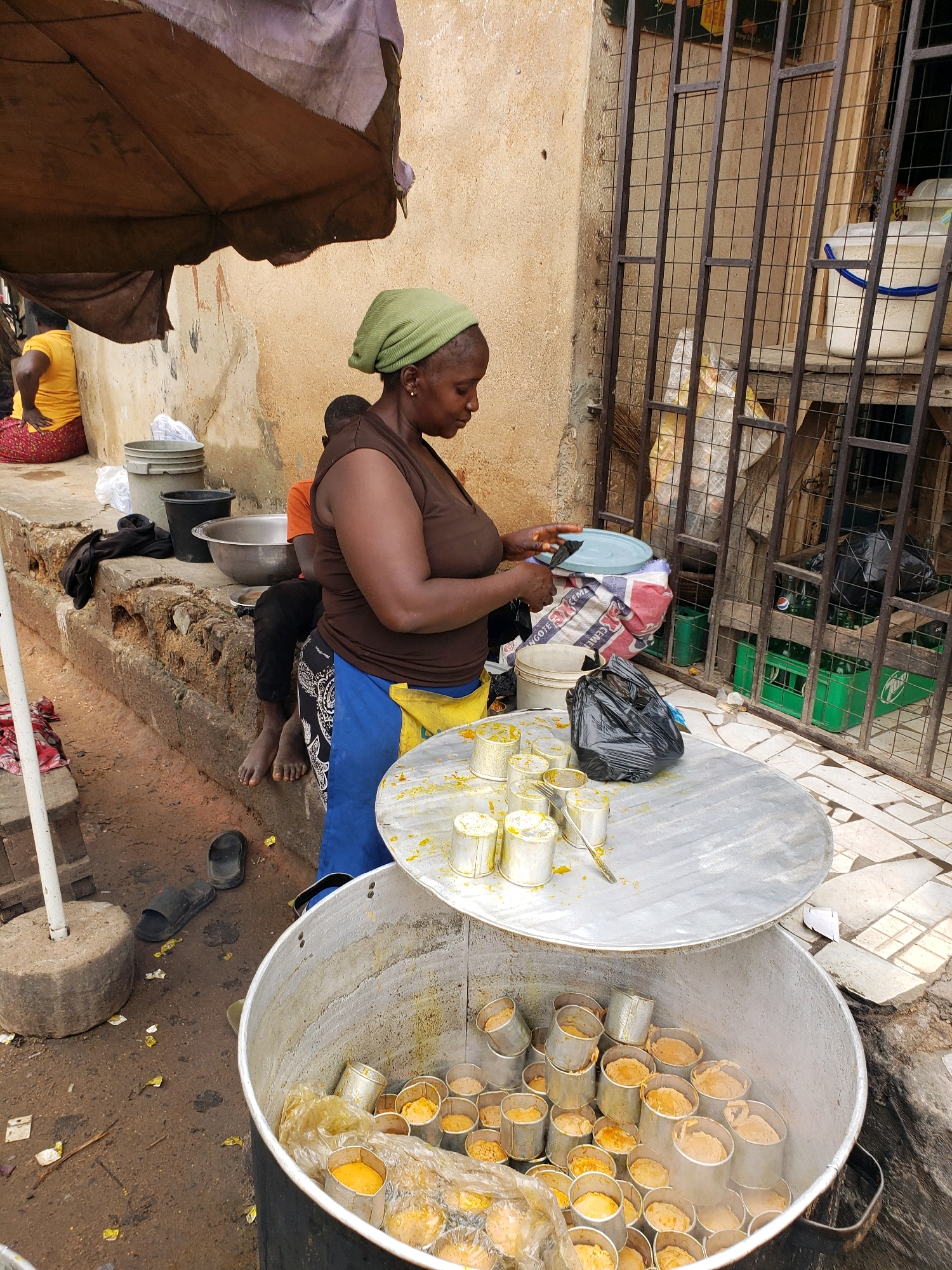 In Narayi, Nigeria, a woman wearing a green headscarf and a brown shirt is preparing food. She is standing beside a large, circular metal container filled with small metal cups, each holding a portion of yellow food. A large metal lid rests on top of the container, and on it, more of these small cups are arranged, some with yellow food inside, and a fork. To her left, there are several buckets, one gray and one black, and a large metal bowl. A child is visible sitting on a raised ledge behind the woman. In the background, a metal gate with a grid pattern encloses a space containing crates of green bottles, possibly beverages. The ground is a mix of dirt and tiled pavement. An umbrella shades part of the scene.