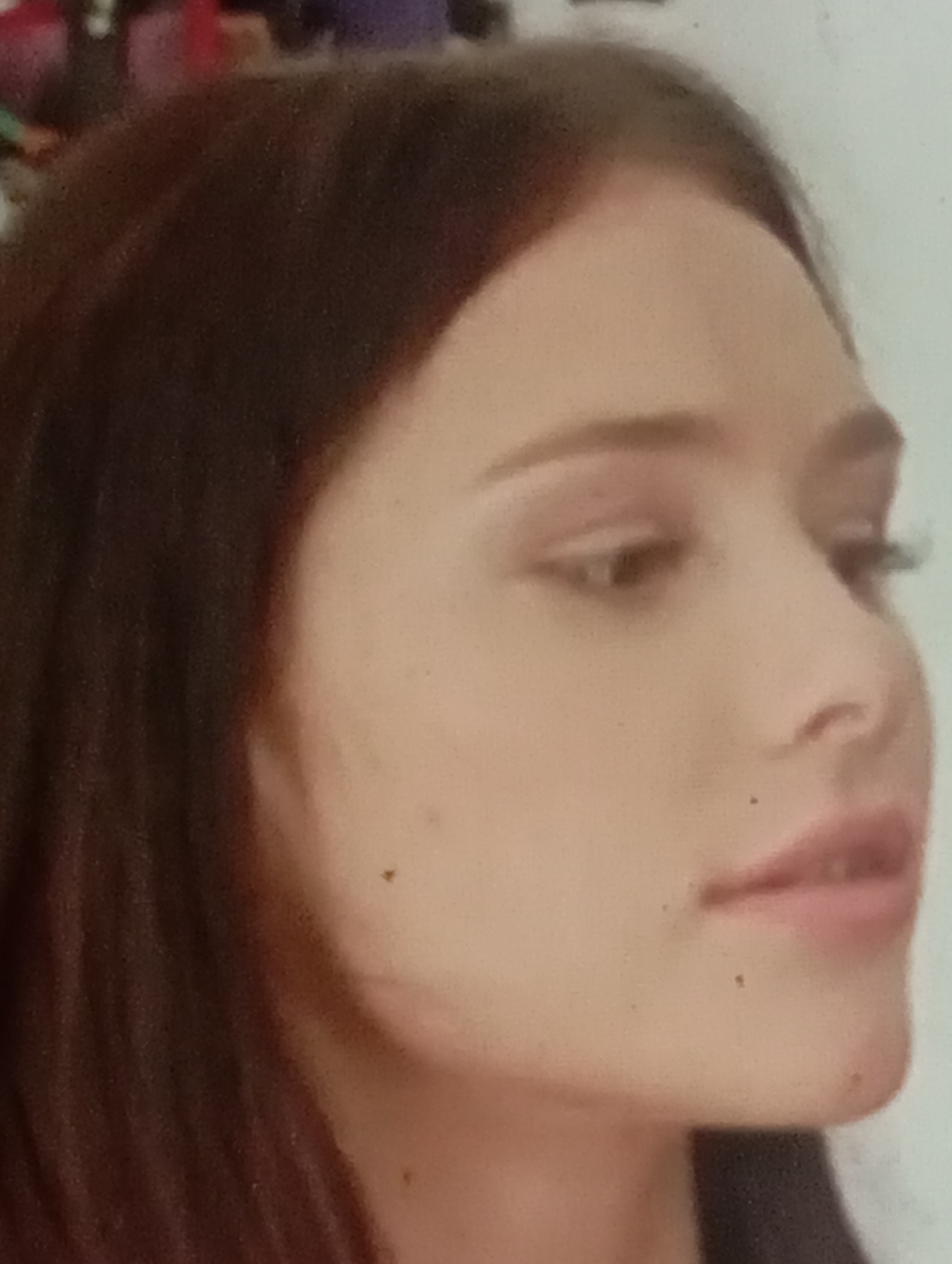 The image is a close-up profile view of a young woman's face, with her head turned to the right. She has dark brown hair that falls around her face and shoulders. Her skin is fair, with a few small dark blemishes visible on her cheek and chin. Her eye is visible, with light brown irises and dark eyelashes. Her lips are slightly parted, revealing a hint of her teeth. The background is out of focus, but appears to be a light-colored wall. The image was taken in Sofiivska Borshchahivka, Ukraine.