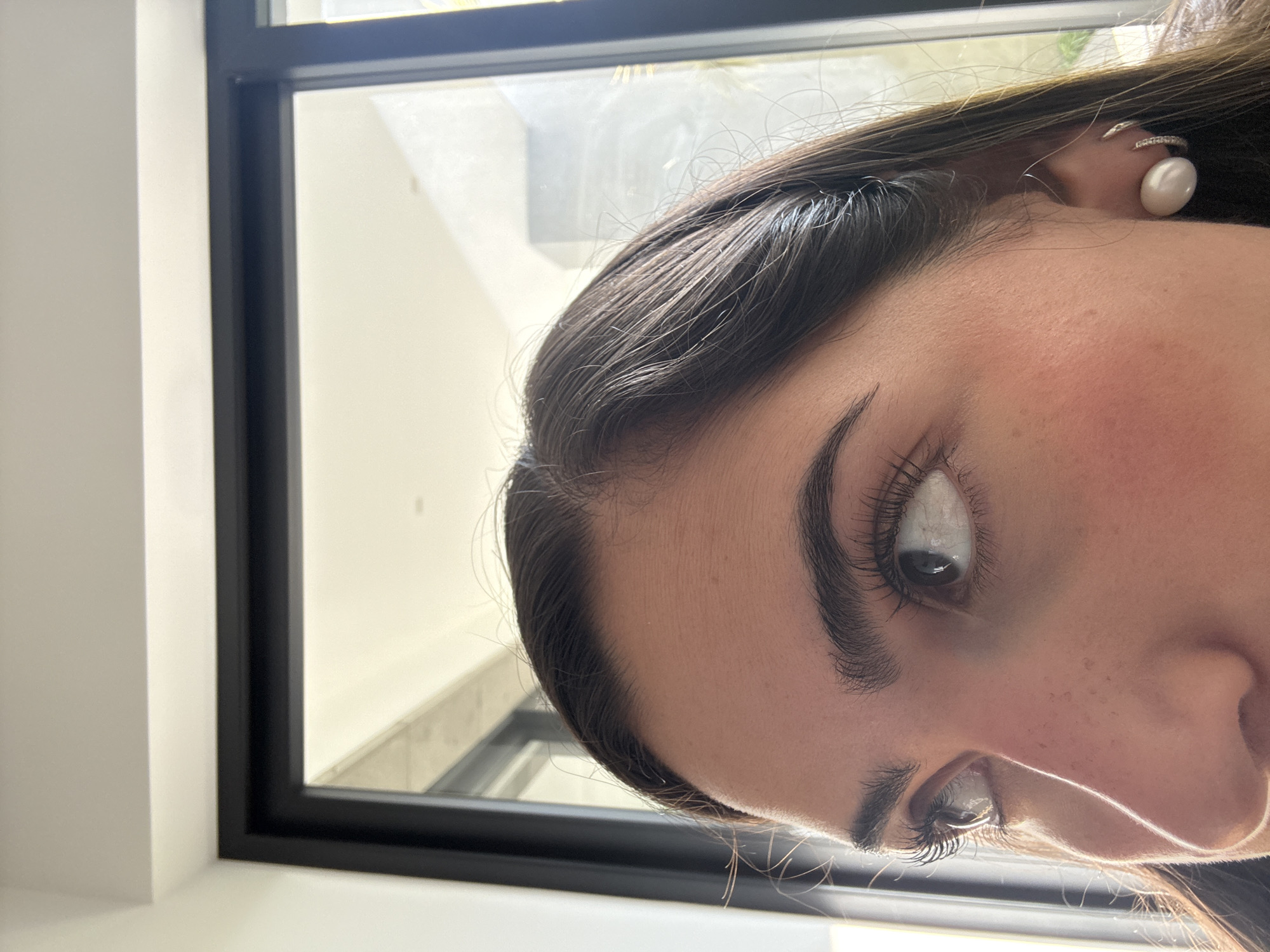 A close-up, tilted shot depicts a person's face from the right side, positioned near a window. The individual has dark hair and visible eyelashes and eyebrows. A pearl earring is visible on the right ear. The background includes a window frame and a glimpse of the exterior. The location in the city of San Bernardino Tlaxcalancingo, Mexico, is not discernible from the image.