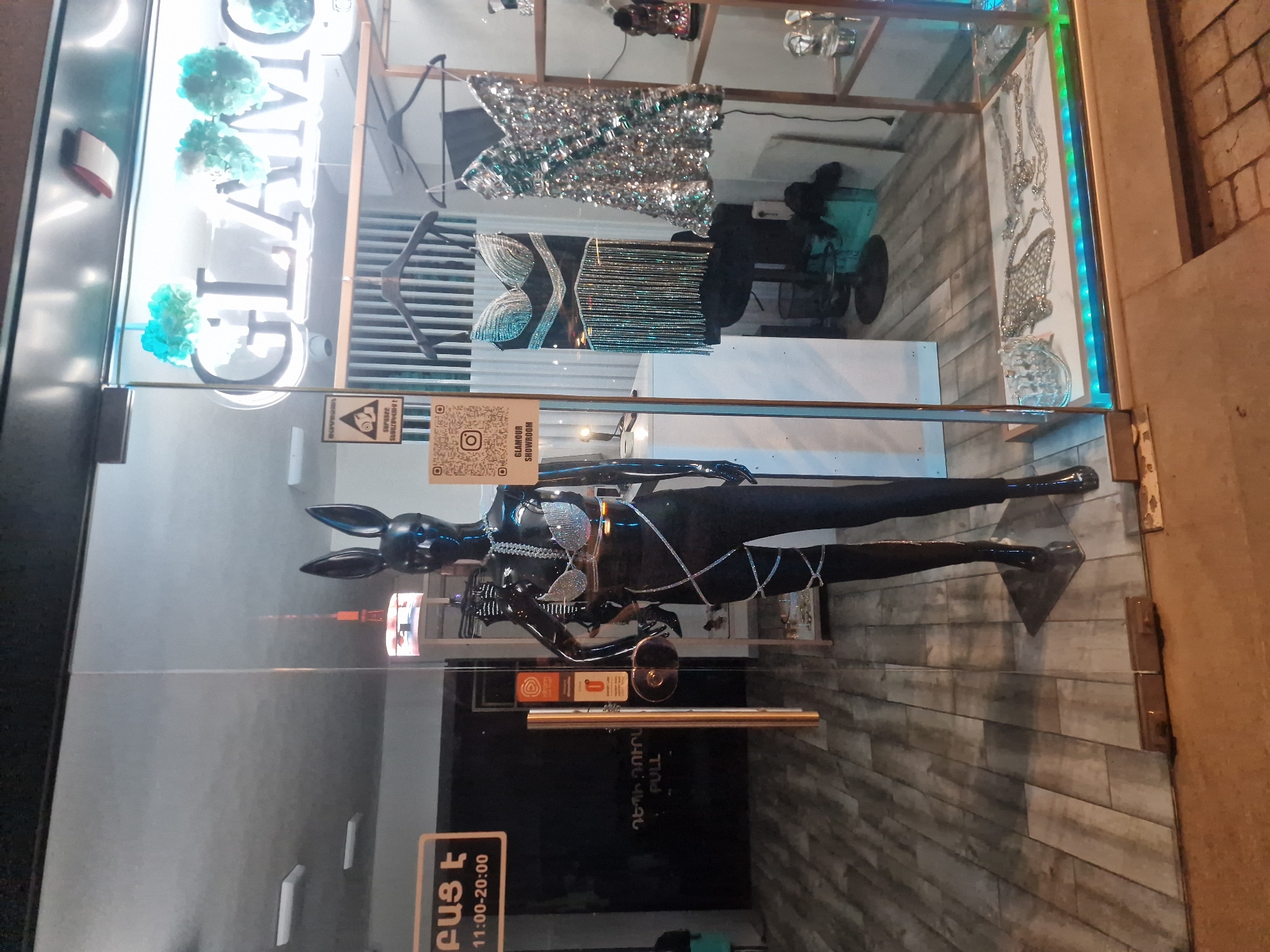 A black mannequin wearing a sparkly bra and black pants with a silver criss-cross design stands in the window of a store named "GLAMOUR SHOWROOM." The store has a glass front, and the mannequin is positioned slightly to the left of center. Behind the mannequin, the store's interior is visible, with clothing racks holding two dresses, one black with fringe and the other silver and sequined. There are also decorative elements like blue and white pom-poms hanging above the "GLAMOUR SHOWROOM" sign. A QR code and an Instagram logo are visible on the left side of the window. The floor of the store appears to be wooden planks. To the left of the mannequin, a sign displays business hours in Armenian and English. The storefront is lit, and the exterior sidewalk is visible in the foreground.