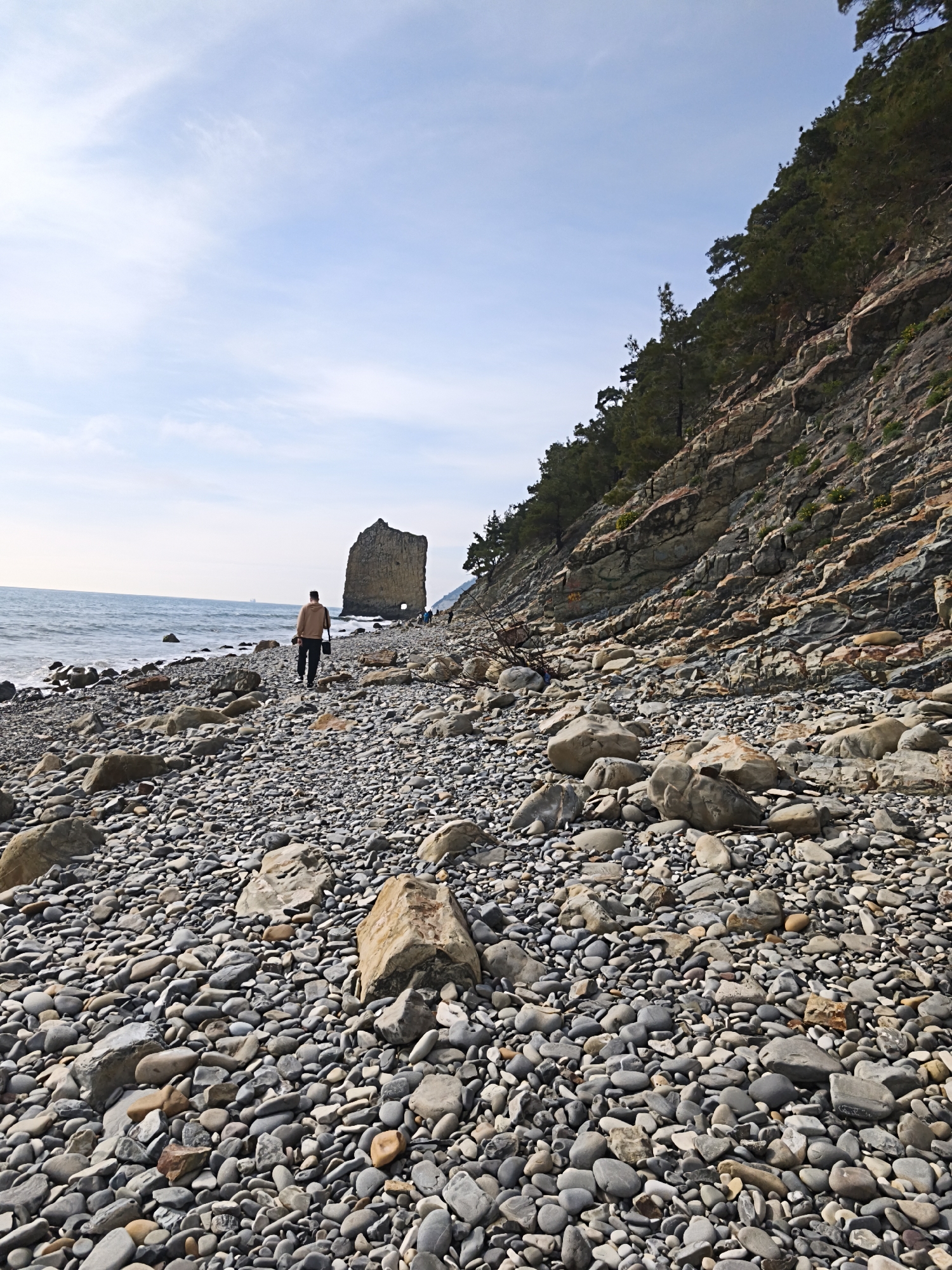 The image captures a rocky beach scene in Praskoveyevka, Russia. A man is walking along the beach, away from the viewer, toward a large, isolated rock formation in the water. The beach is covered in various sizes of smooth, grey rocks. To the right, a steep, rocky cliff slopes upward, covered with trees. The sea is visible to the left, with gentle waves. The sky is light blue with some scattered clouds.