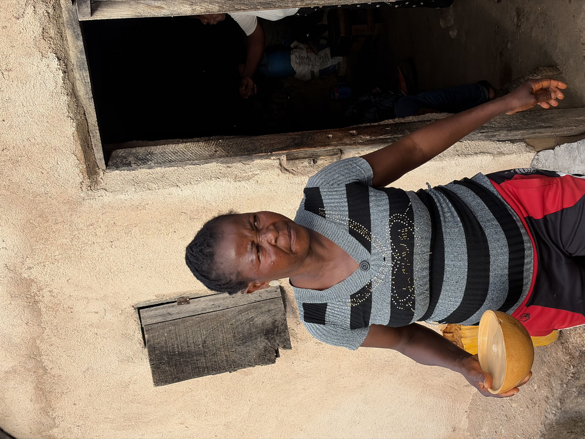 This image, captured in the city of Jos, Nigeria, displays an adult female standing outdoors against a textured, light-beige building facade. She has dark skin and short, dark hair styled back. Her facial expression is focused, with a slight grimace, as she looks towards the right side of the frame. She wears a short-sleeved V-neck top with horizontal stripes of light grey and black, the black stripes embellished with small white dots. Her lower garment consists of black athletic shorts featuring a red panel on the upper left thigh. Gold hoop earrings are visible. In her left hand, she holds a light brown, organic-material bowl containing a light-colored liquid. To her left, a small, weathered wooden window shutter is partially open within the wall. To her right, an open, rustic wooden door frame leads into a dark interior. Inside this dimly lit space, the upper torso of another individual, wearing a white shirt and dark lower garment, is partially visible, with both hands raised as if performing an action. Identifiable objects within the interior include a blue plastic drum and a white sack bearing partial black text. The immediate ground surface appears rough and earthy.