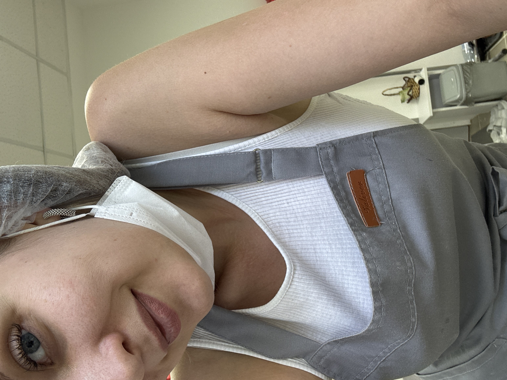 A person wearing a hairnet, a white ribbed tank top, a grey apron with a brown label, and a white face mask is looking at the camera. The background shows white tiled walls and what appears to be kitchen equipment. The location is likely indoors, possibly in a kitchen or food preparation area. There is no information available in the image to ascertain this photo was taken in Izhevsk, Russia.