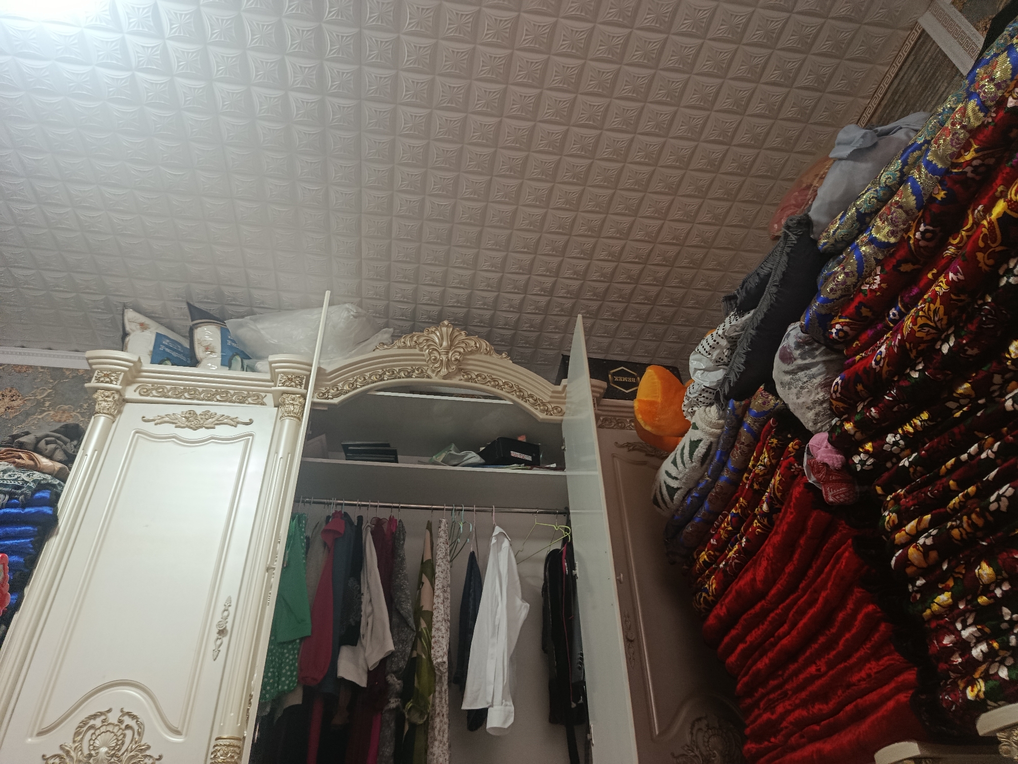 This is a photograph of the interior of a room, likely a closet or dressing room, located in Üç-Korgon, Kyrgyzstan. The scene is dominated by a large, ornate white wardrobe with gold detailing. The wardrobe's left door is closed and features decorative carvings. The right side of the wardrobe is open, revealing a closet interior with a hanging rack filled with various items of clothing. Colors observed in the clothing include green, red, white, and black. The closet shelves above the hanging clothes are sparsely populated with items, including what appear to be books or folders and a black box.
To the right of the wardrobe, a stack of brightly colored textiles, possibly blankets or rugs, are piled high. These textiles display intricate patterns in red, blue, gold, and yellow. A bright orange spherical object is also visible among the textiles.
The ceiling is textured with a repeating geometric pattern, common in interior design. The overall impression is one of a well-organized but somewhat cluttered space, with a focus on textiles and storage. No people are visible in the image.