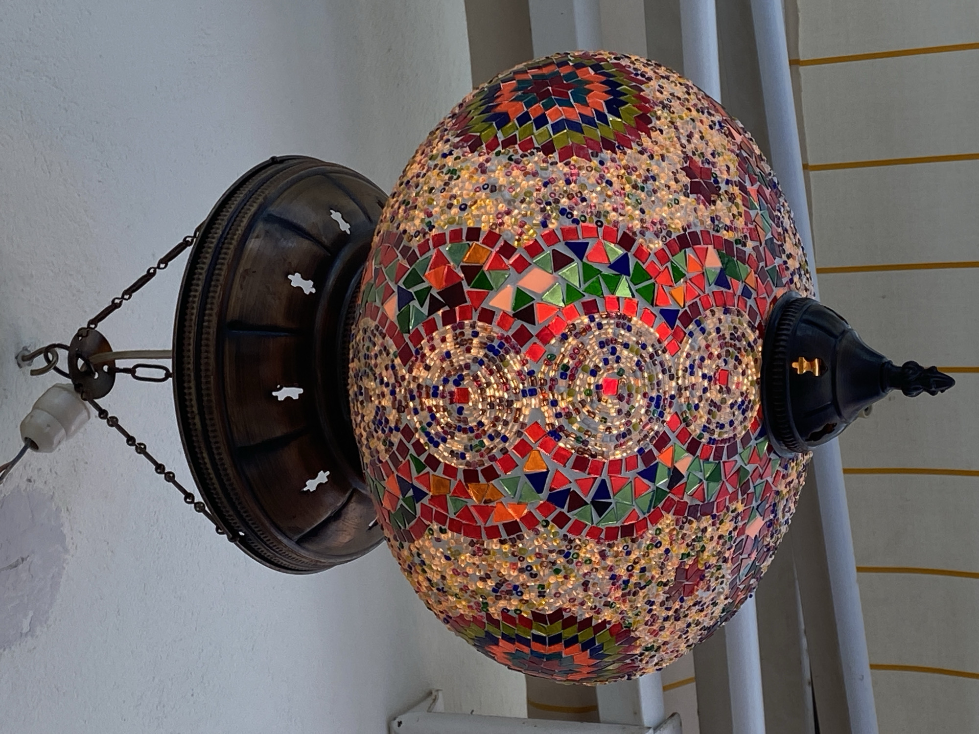 A close-up shot depicts a lit Turkish mosaic lamp hanging from a wall in what is likely a building in Alanya, Turkey. The lamp, predominantly oval in shape, displays a vibrant mosaic pattern of small colorful pieces, including reds, oranges, greens, blues, and yellows. At the top, larger, more defined squares and triangles create a dense mosaic of colors, while the lower portion features a more densely packed, speckled pattern. A circular motif is visible in the center, with concentric rings of red and other colors.
The lamp is connected to the wall by a dark, bronze-colored metal fixture that resembles a shallow bowl with decorative cutouts. A chain connects this fixture to the electrical wiring. The lamp is illuminated, causing the colored pieces to glow. The light source is situated inside the lamp. The background includes plain white walls. To the right of the lamp are vertical lines of a light, off-white/tan color. The scene is indoors and the focus is strongly on the lamp and its construction.